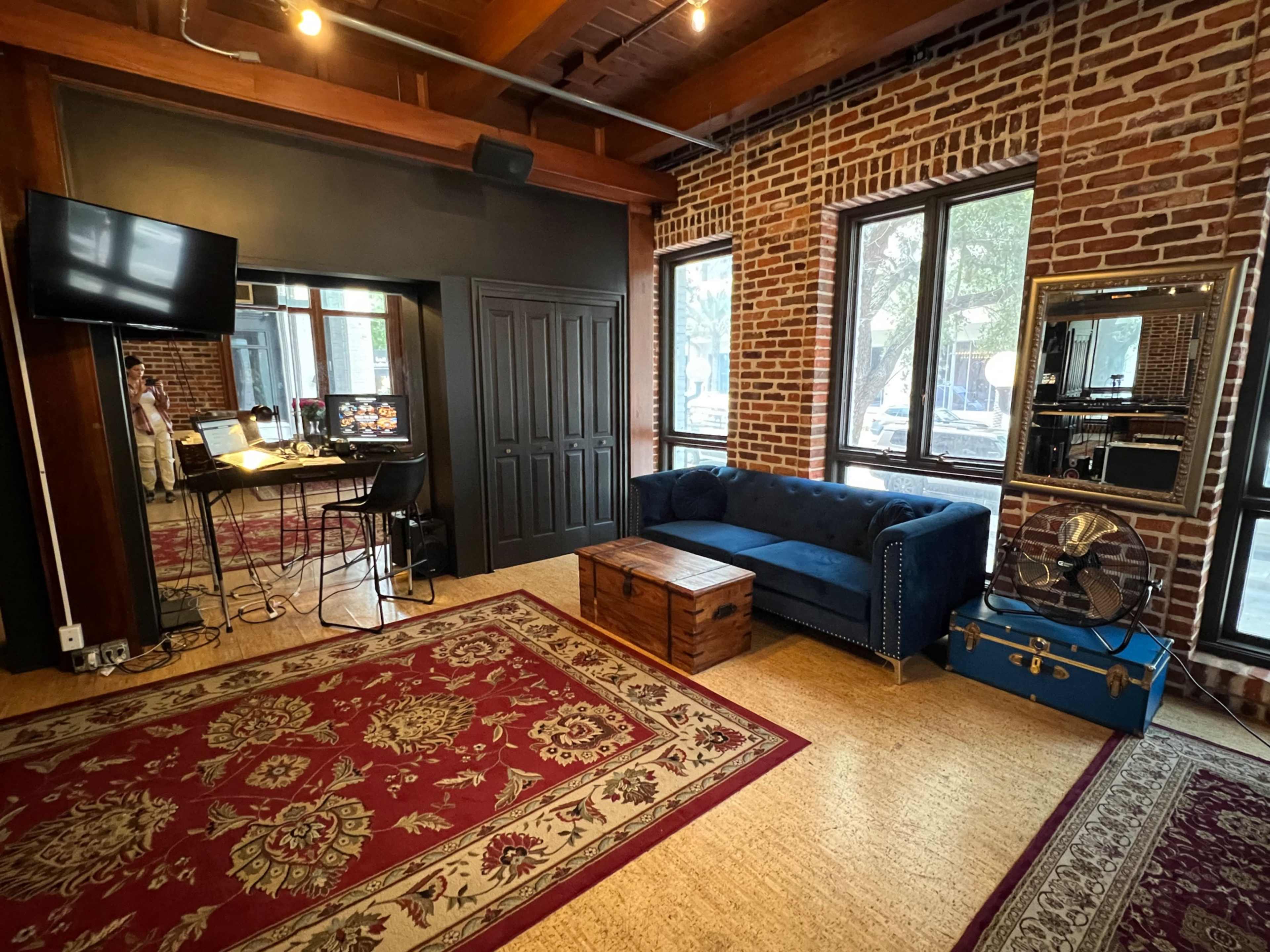 The image shows a room with exposed brick walls, featuring a blue couch, a wooden coffee table, a small desk with a computer, and a large mirror, all complemented by a decorative rug.
