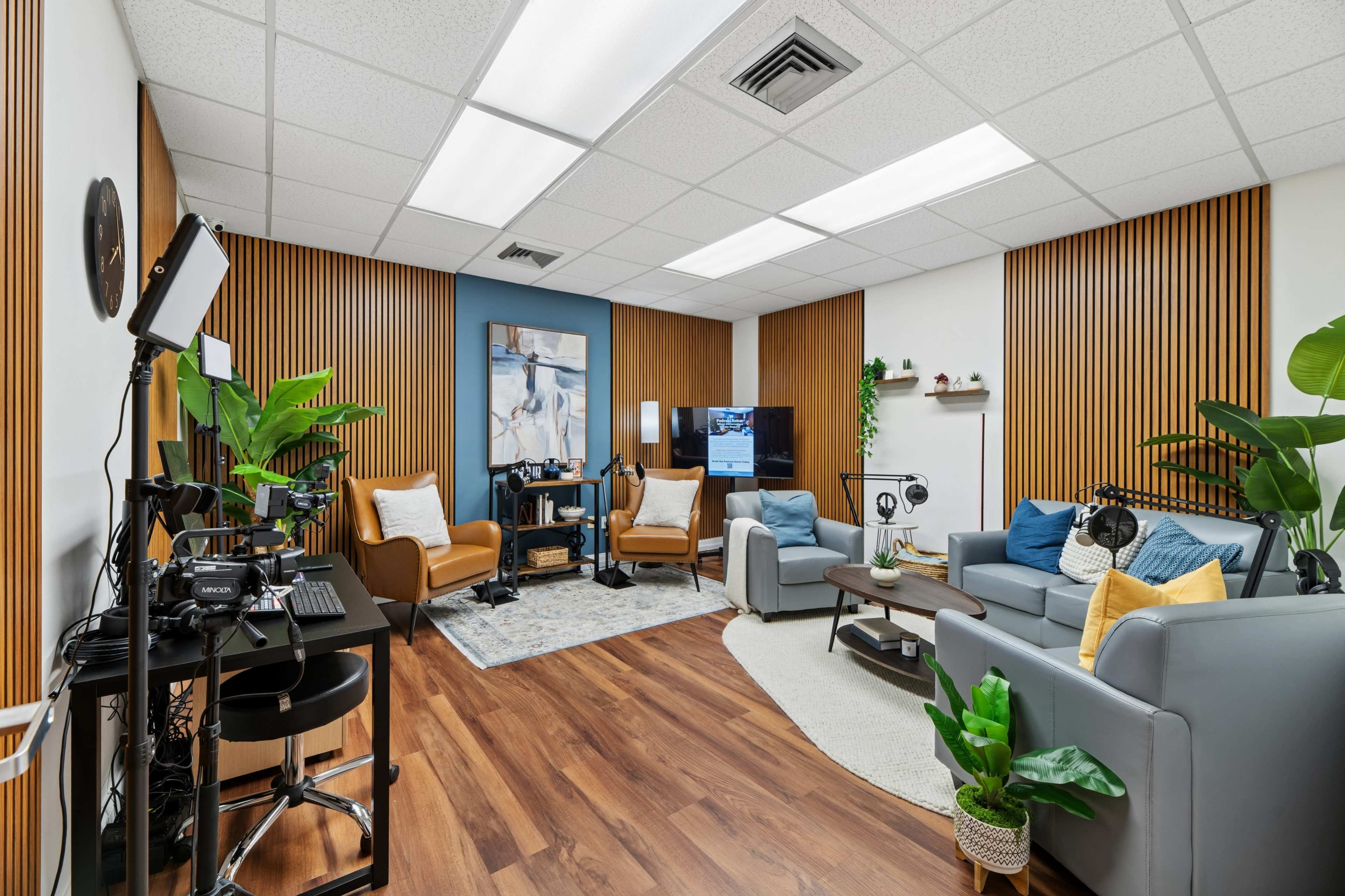 The image shows a well-furnished office space featuring two seating areas, each with multiple chairs, a television, and various plants.