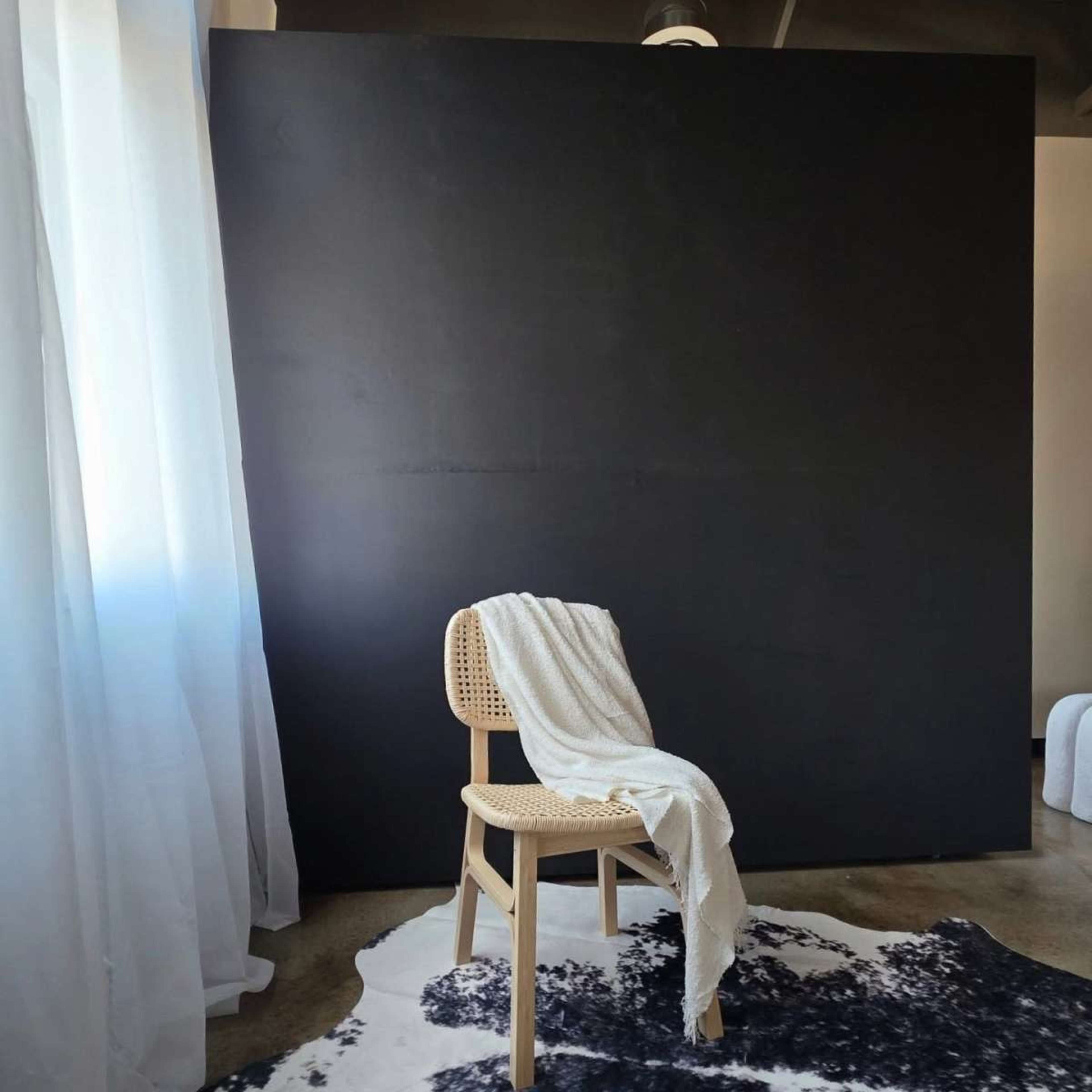 A wooden chair with a blanket draped over it is positioned in front of a large black wall, on a patterned rug, with sheer white curtains nearby.