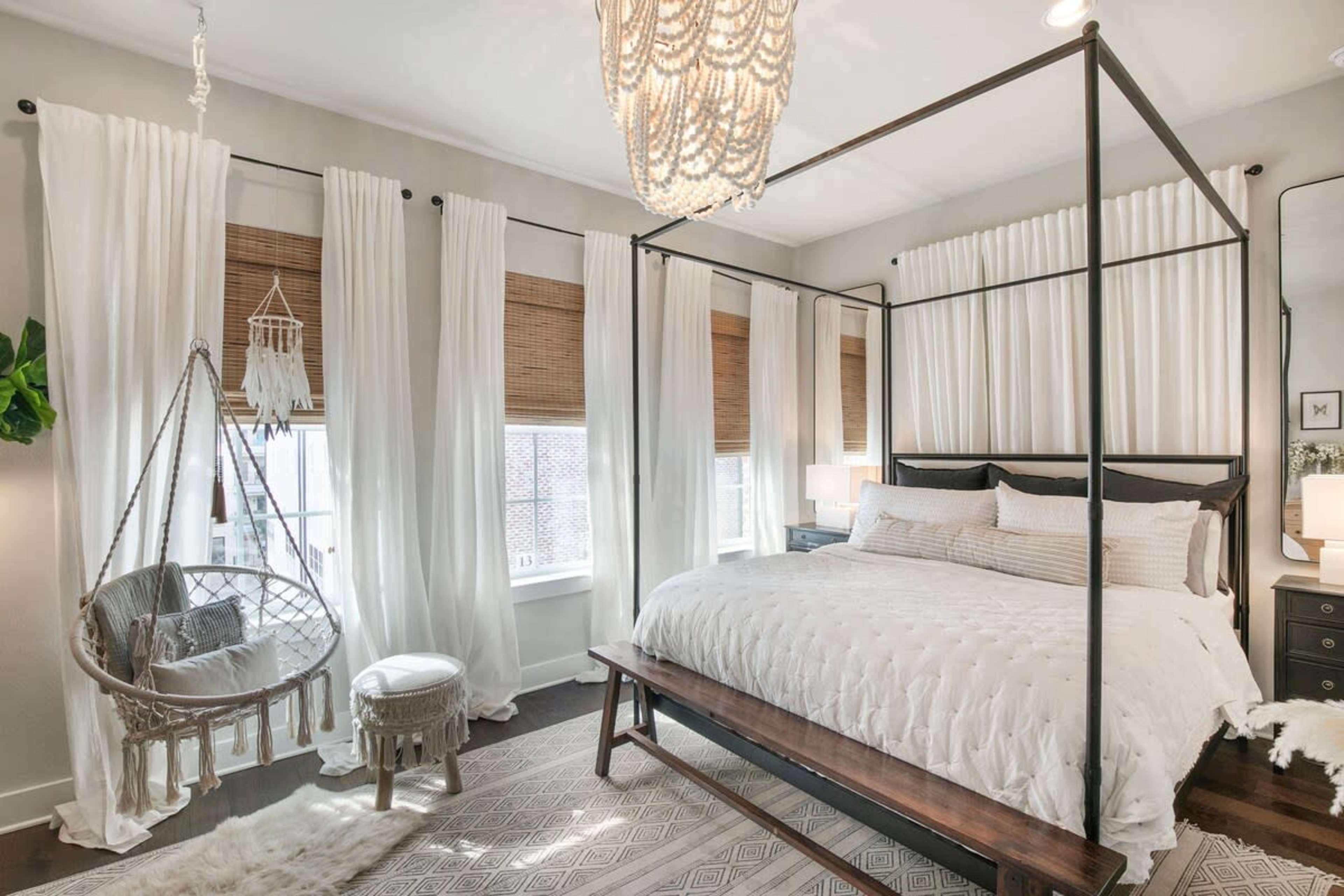 The image shows a bedroom featuring a four-poster bed with white bedding, large windows with bamboo shades, and a hanging chair in the corner.