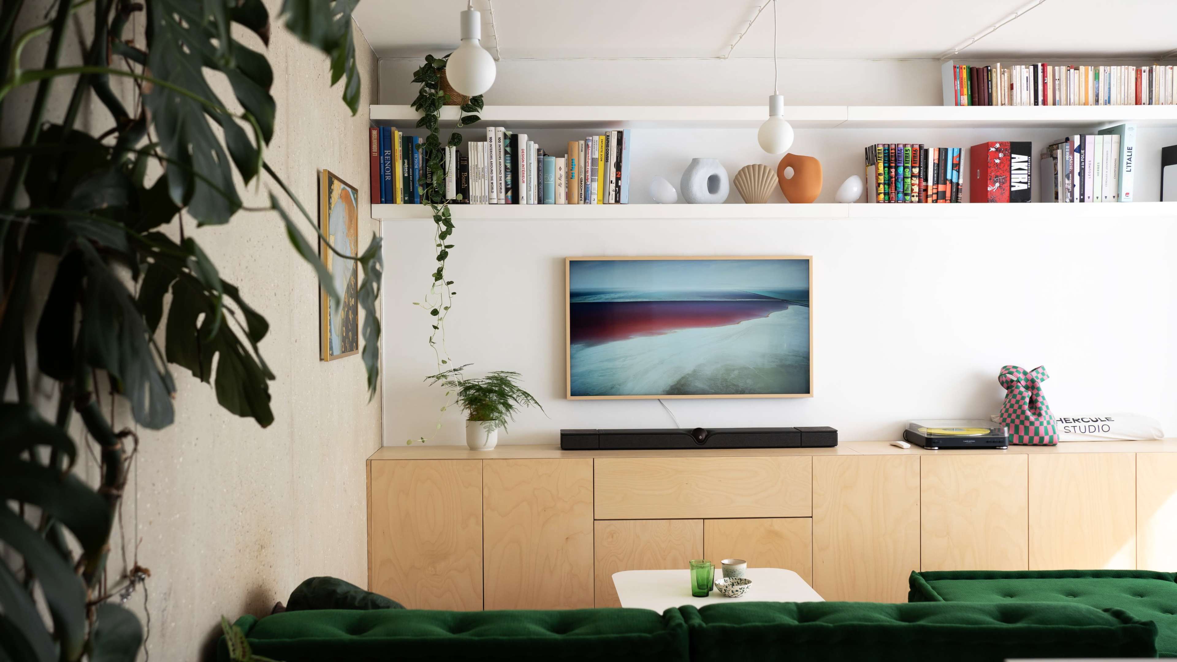 The image shows a modern living room with a green couch, a wooden cabinet, and bookshelves filled with books and decorative items.