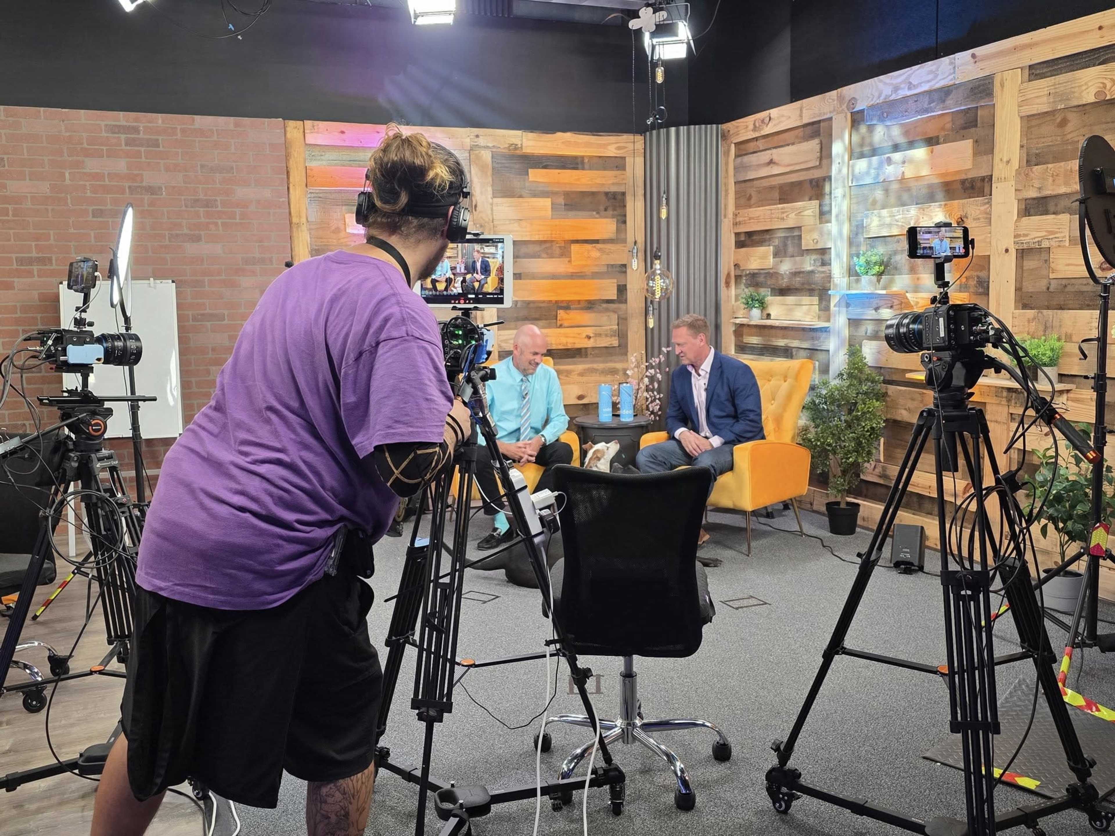 A camera crew records a conversation between two individuals seated in an orange armchair in a well-lit studio with wooden panels and plants.