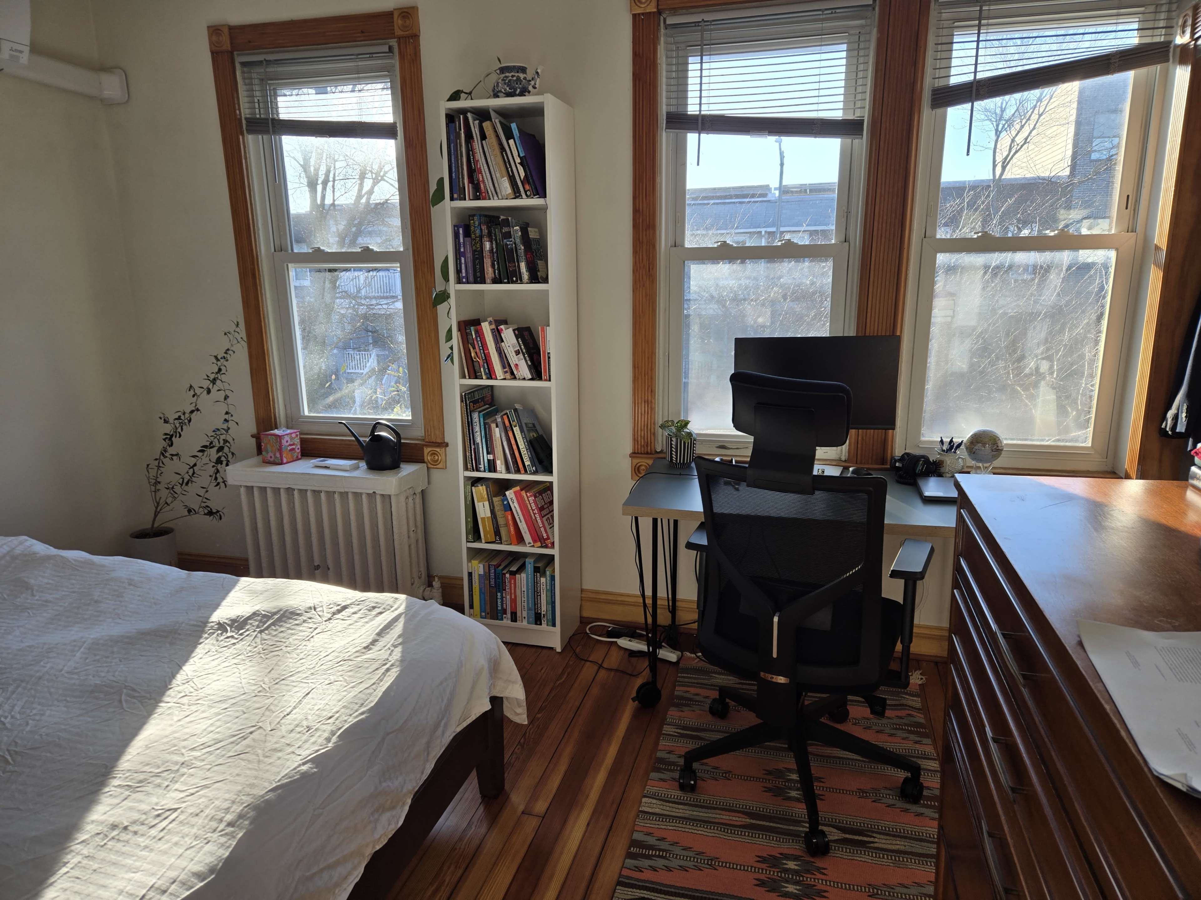 A sunlit room features a bed, a desk with a computer chair, a bookshelf filled with books, and large windows providing a view.