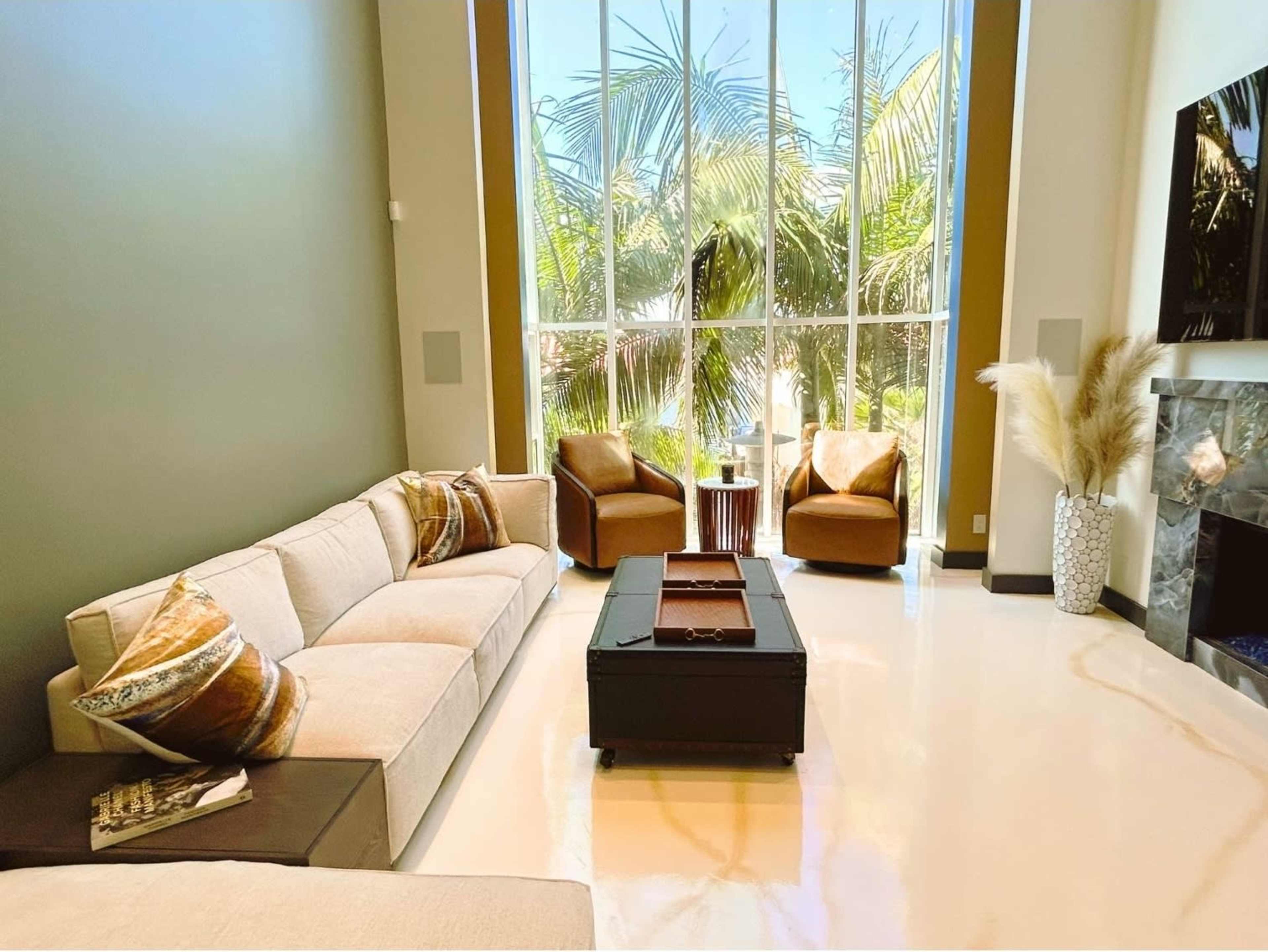 The image shows a modern living room with a large window overlooking palm trees, two armchairs, a beige sofa, and a coffee table.