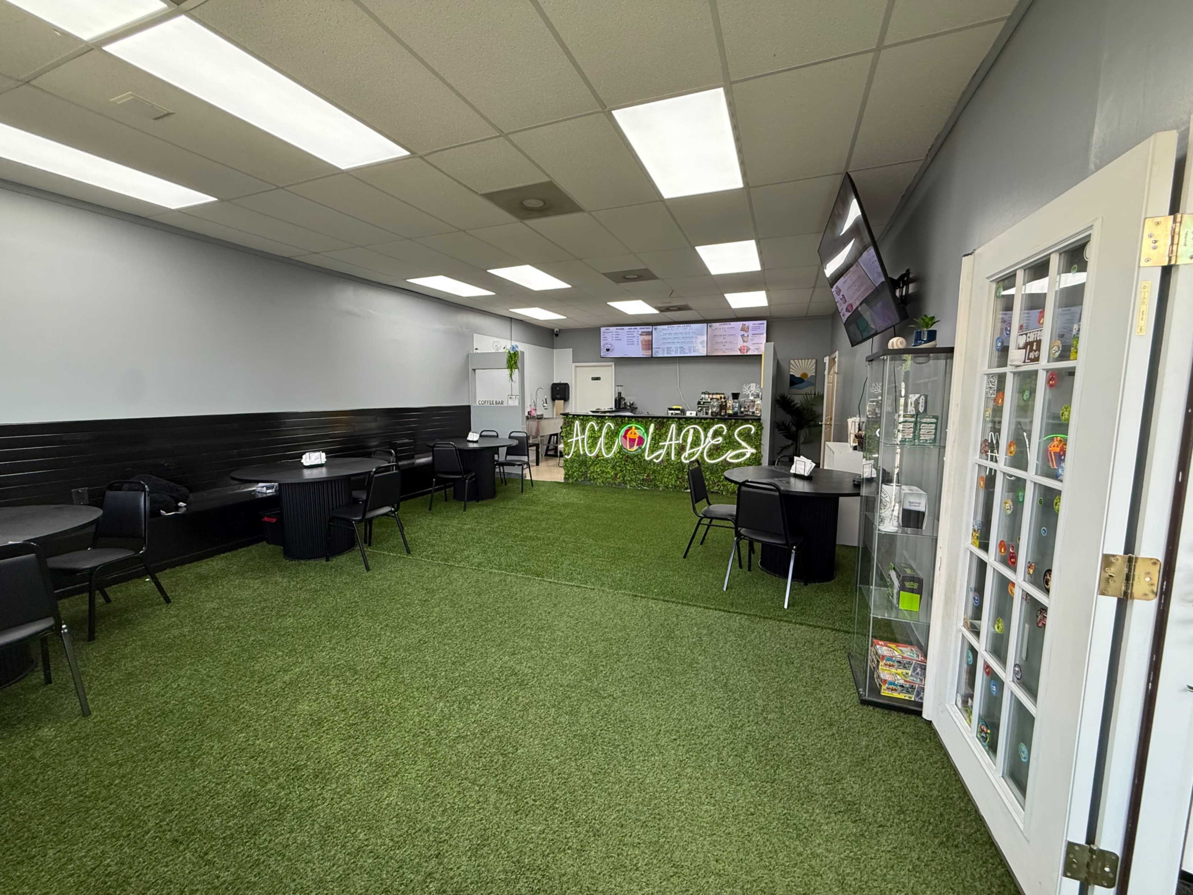 The image shows an interior of a dining area with synthetic grass flooring, tables, and a counter with a menu board.