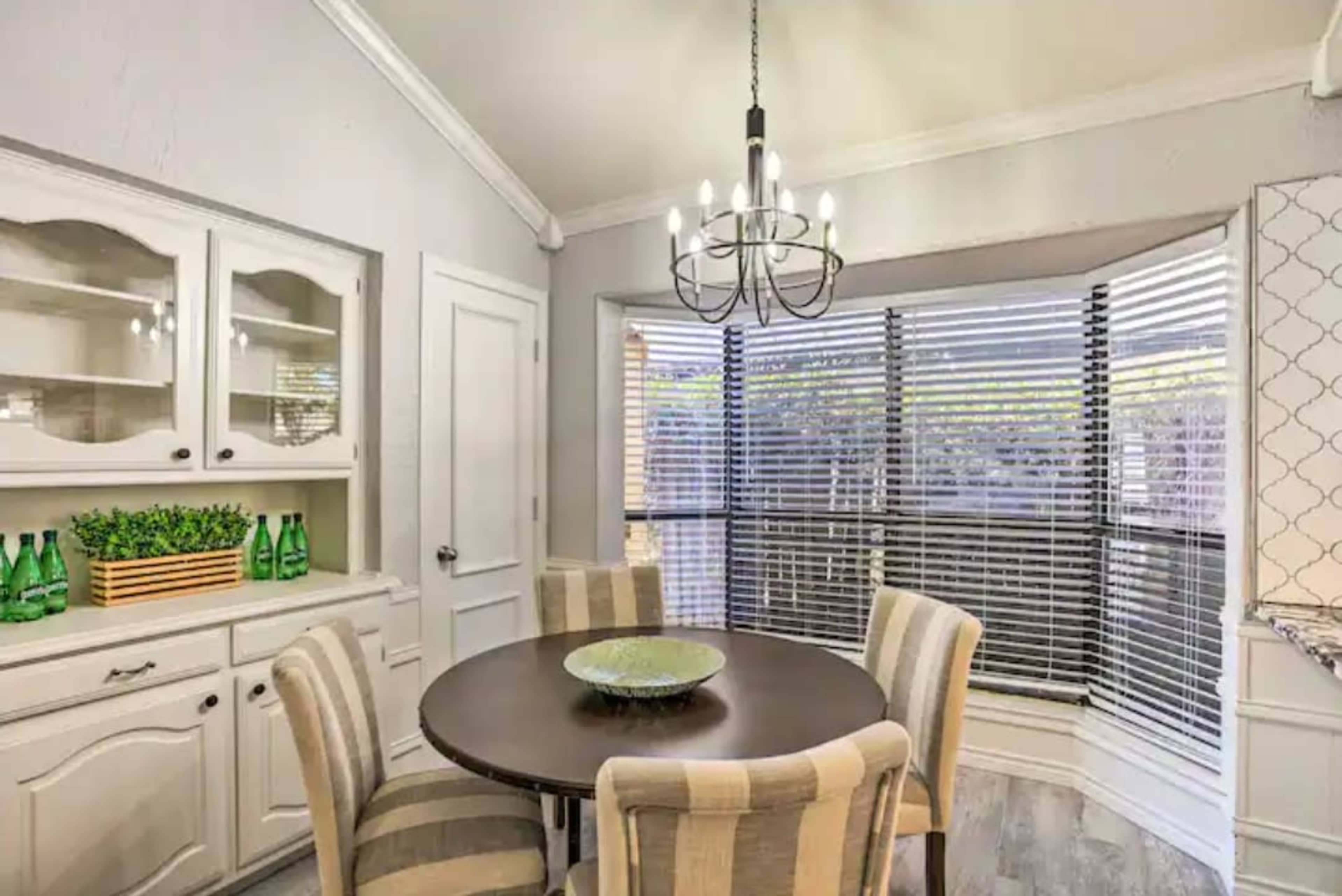 A dining area features a round table surrounded by four striped chairs, with a chandelier overhead and a window with blinds letting in light.