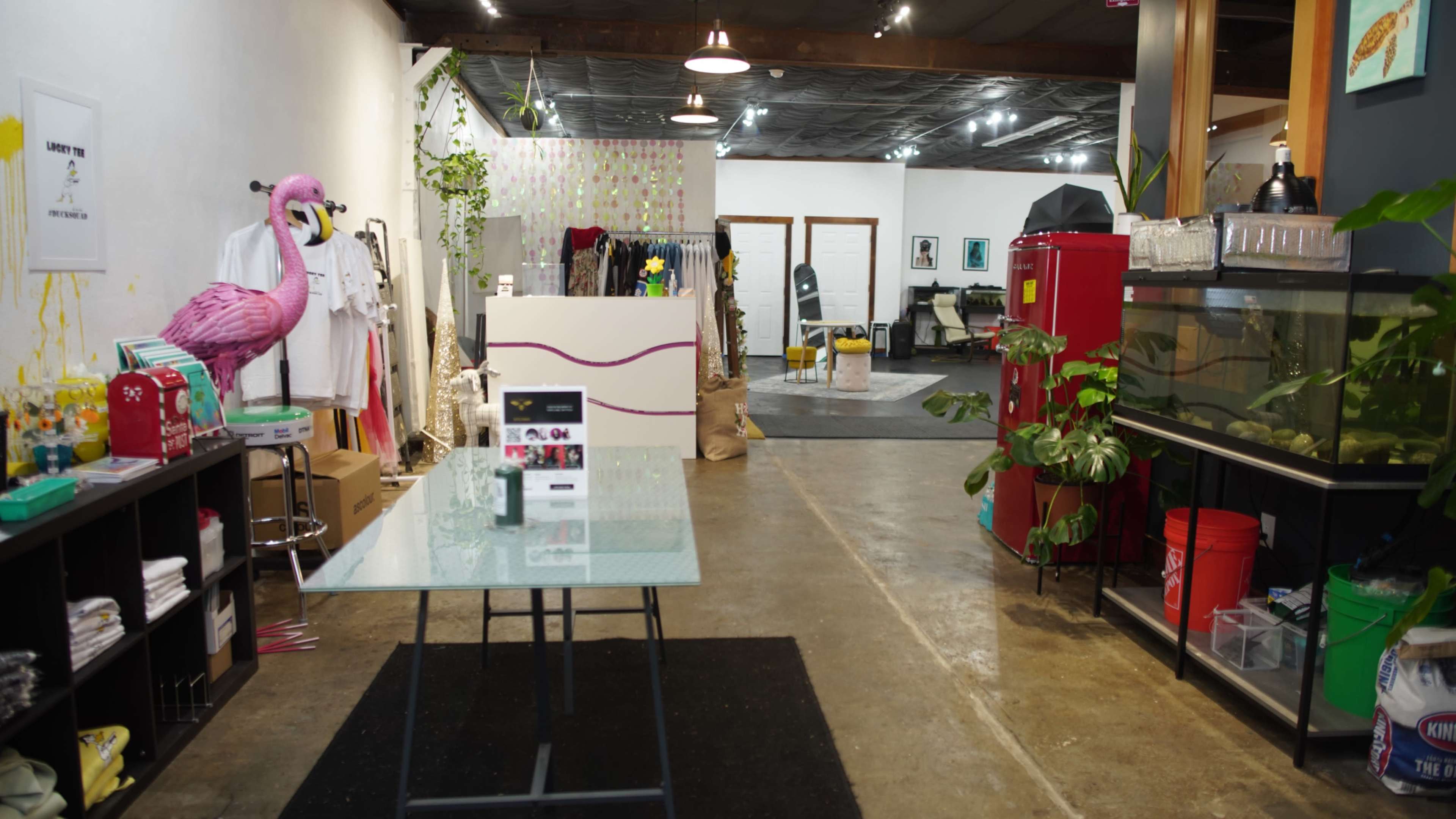 The image shows a retail store interior with clothing racks on one side, a glass table in the foreground, and a seating area in the background.