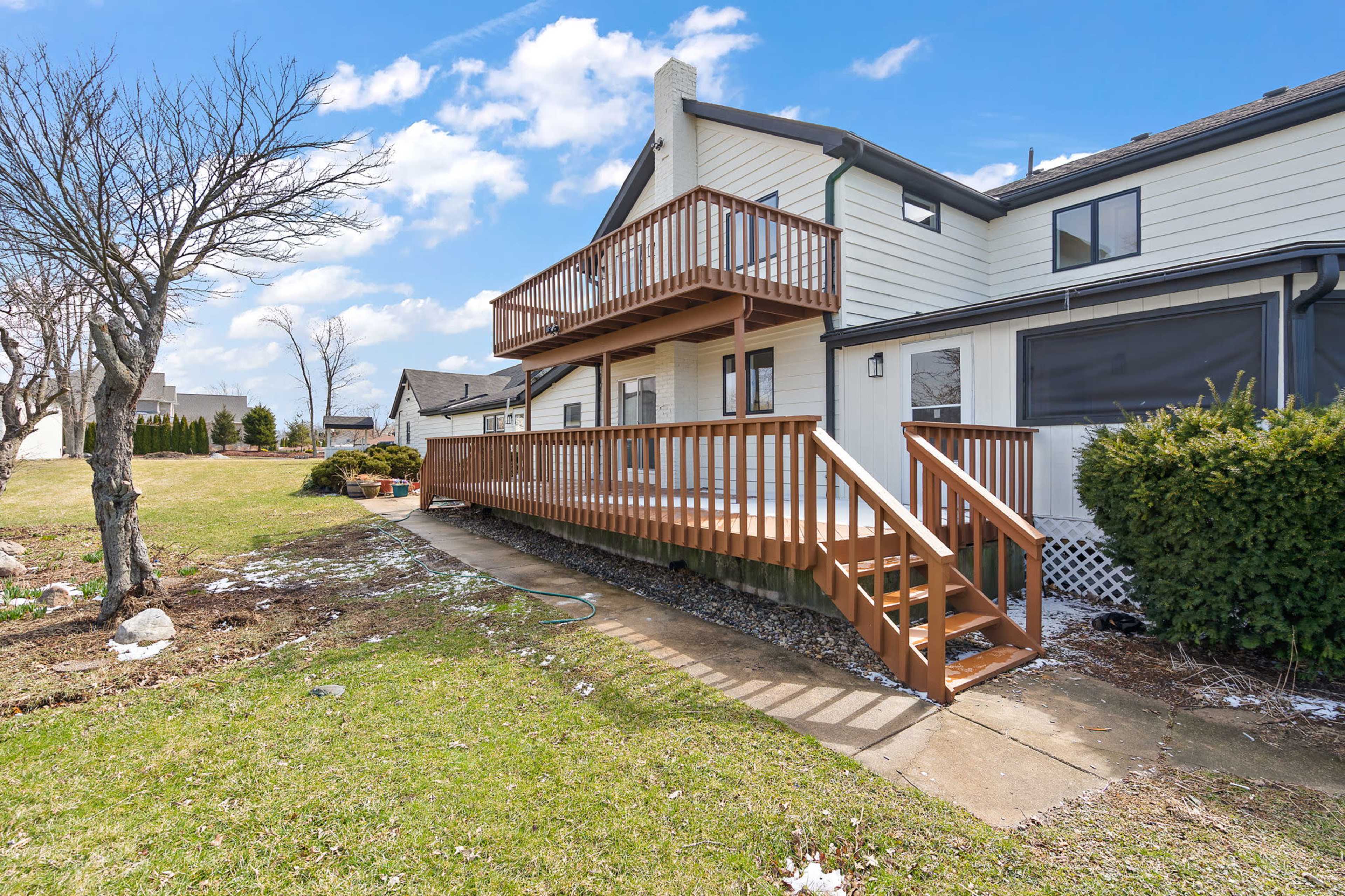 The image shows a house with a wooden deck and railing, surrounded by a grassy yard and a few scattered patches of snow.