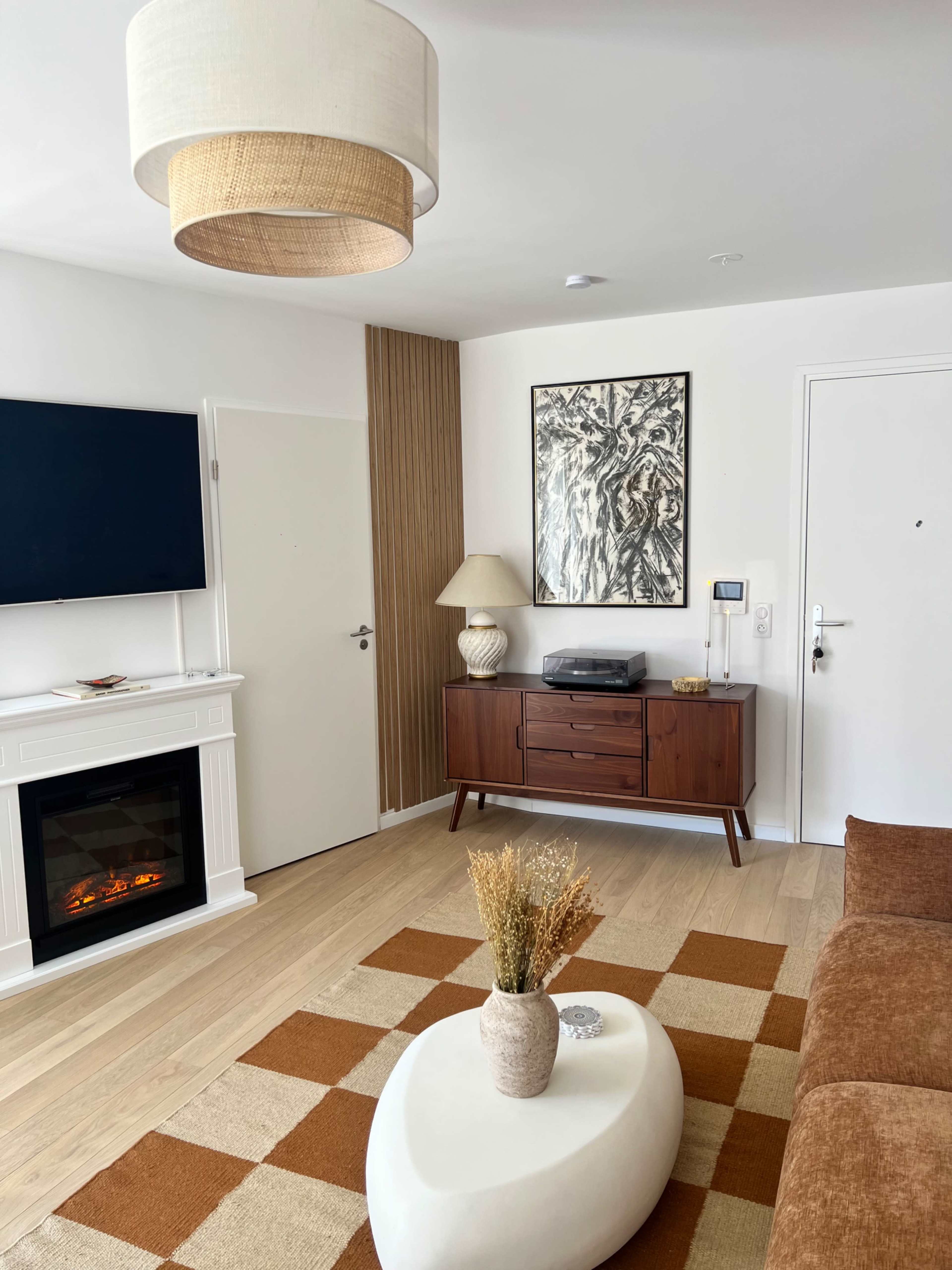 The image shows a stylish living room with a wooden cabinet, a wall-mounted TV, an electric fireplace, and a light-colored coffee table with a decorative vase on a patterned area rug.