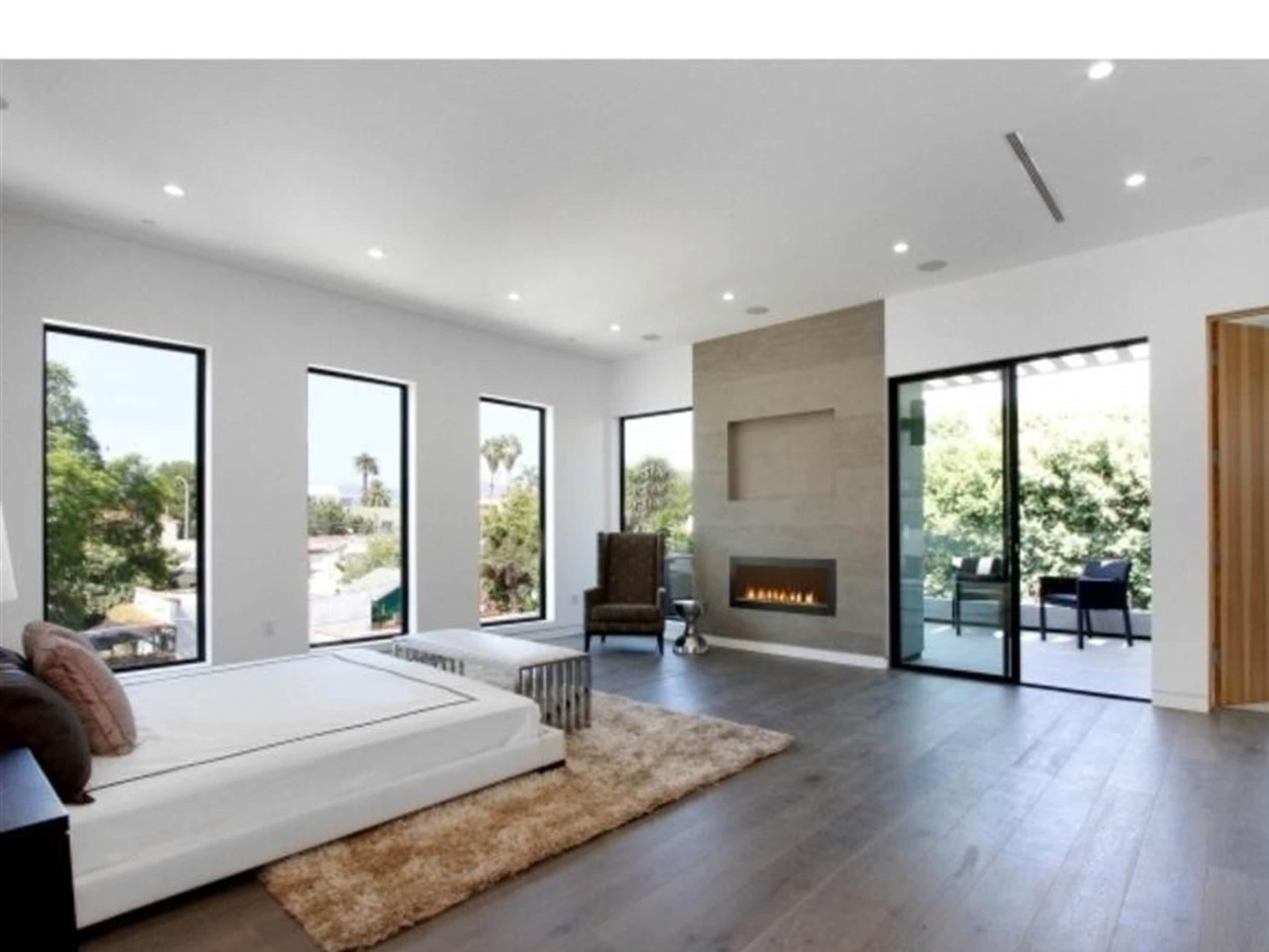 A spacious, modern bedroom with large windows, a minimalist fireplace, and an area rug on a wooden floor.