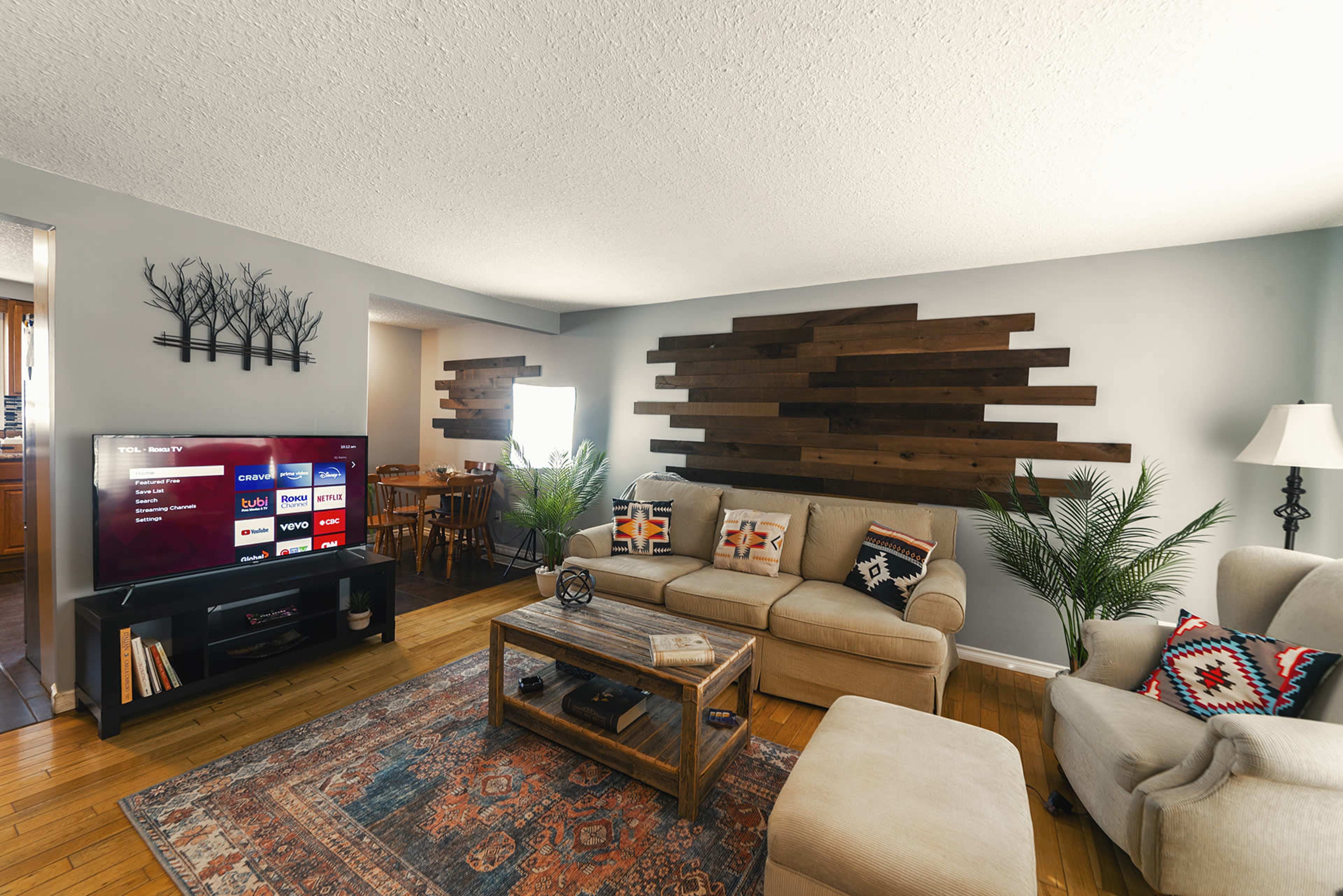 A living room with a beige sofa, an entertainment center with a TV, a wooden coffee table, and decorative plants, set against a backdrop of wooden wall panels.
