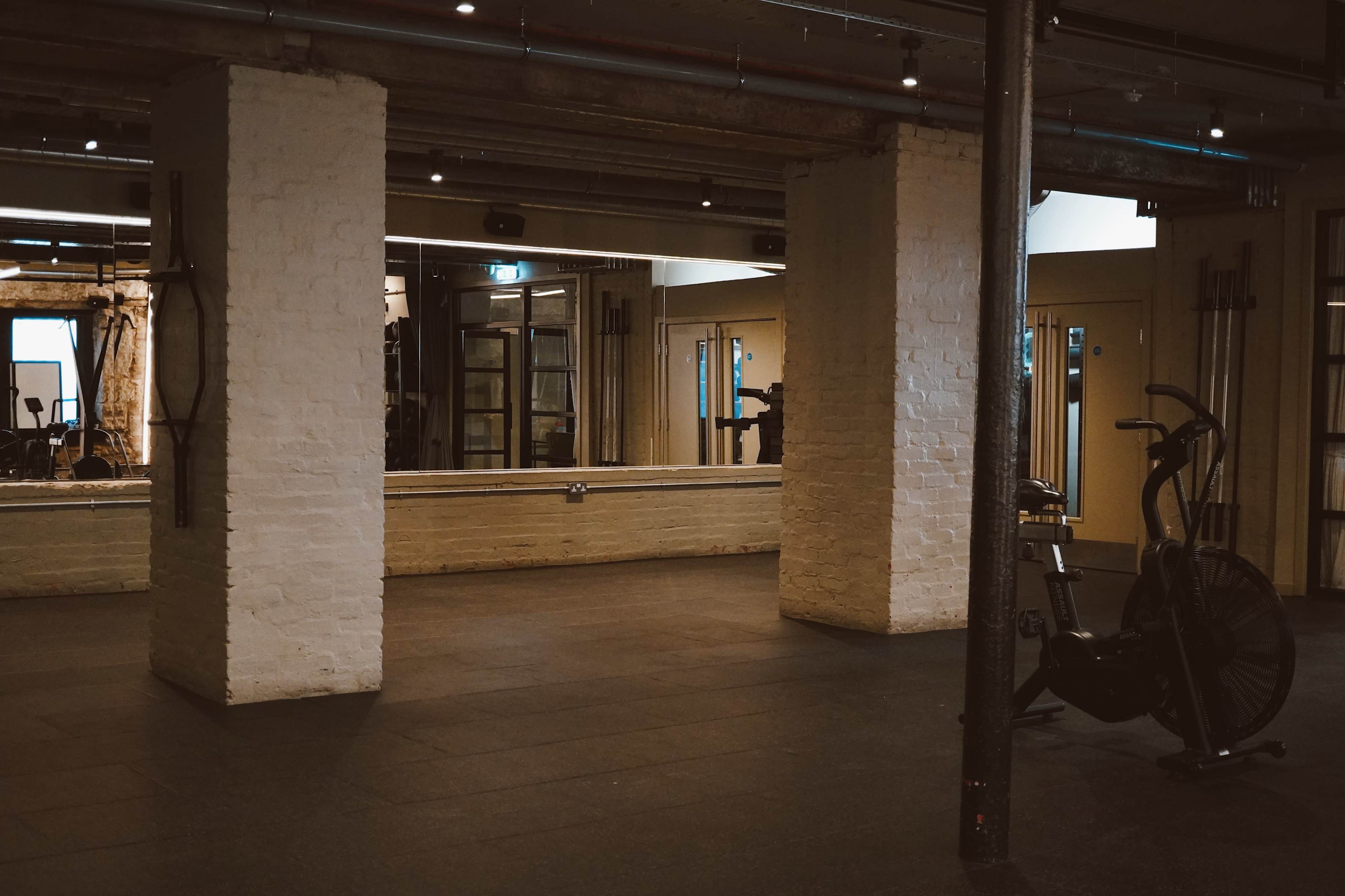 The image shows a spacious, modern gym with exposed brick walls, mirrors, and an exercise bike in the corner.