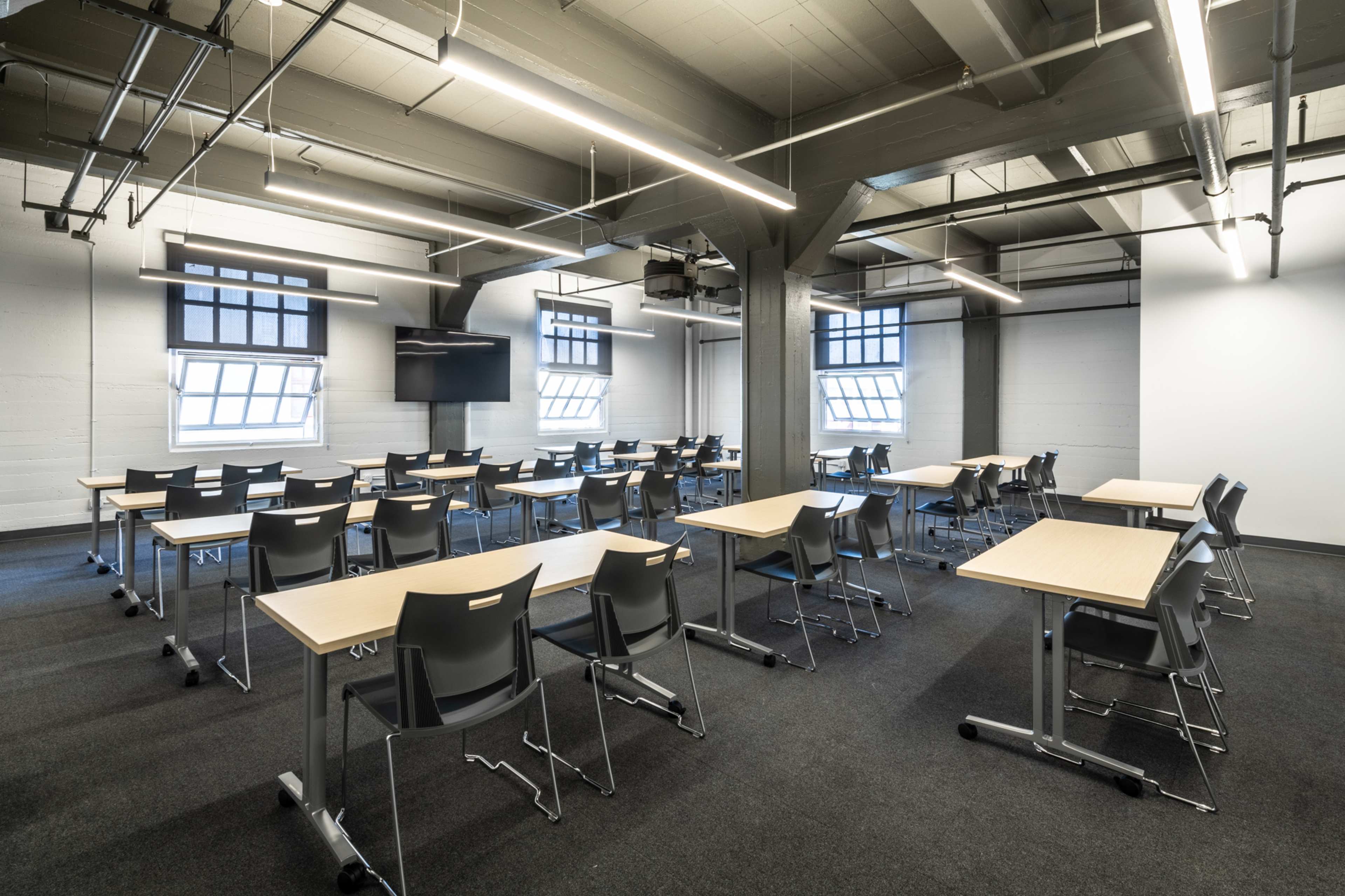 A modern classroom arranged with several tables and chairs facing a wall-mounted screen.