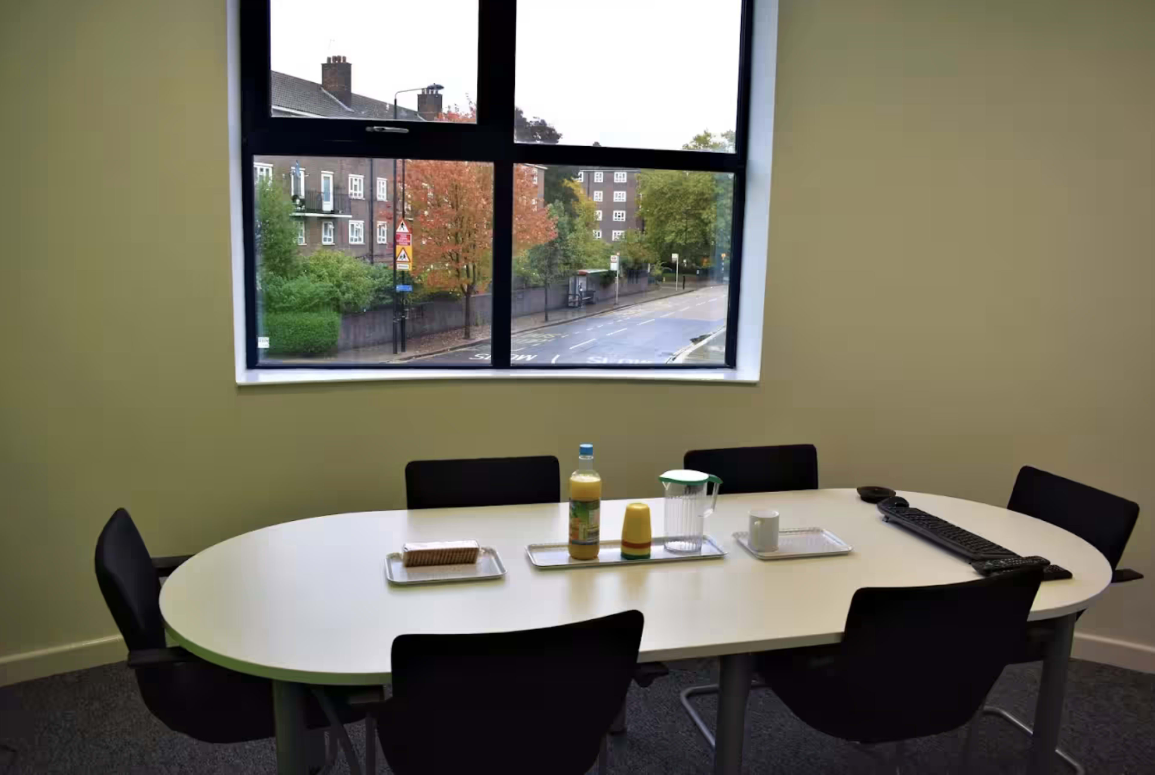 A conference room features a white oval table set with drinks and a laptop, alongside a large window overlooking a street with trees in autumn colors.