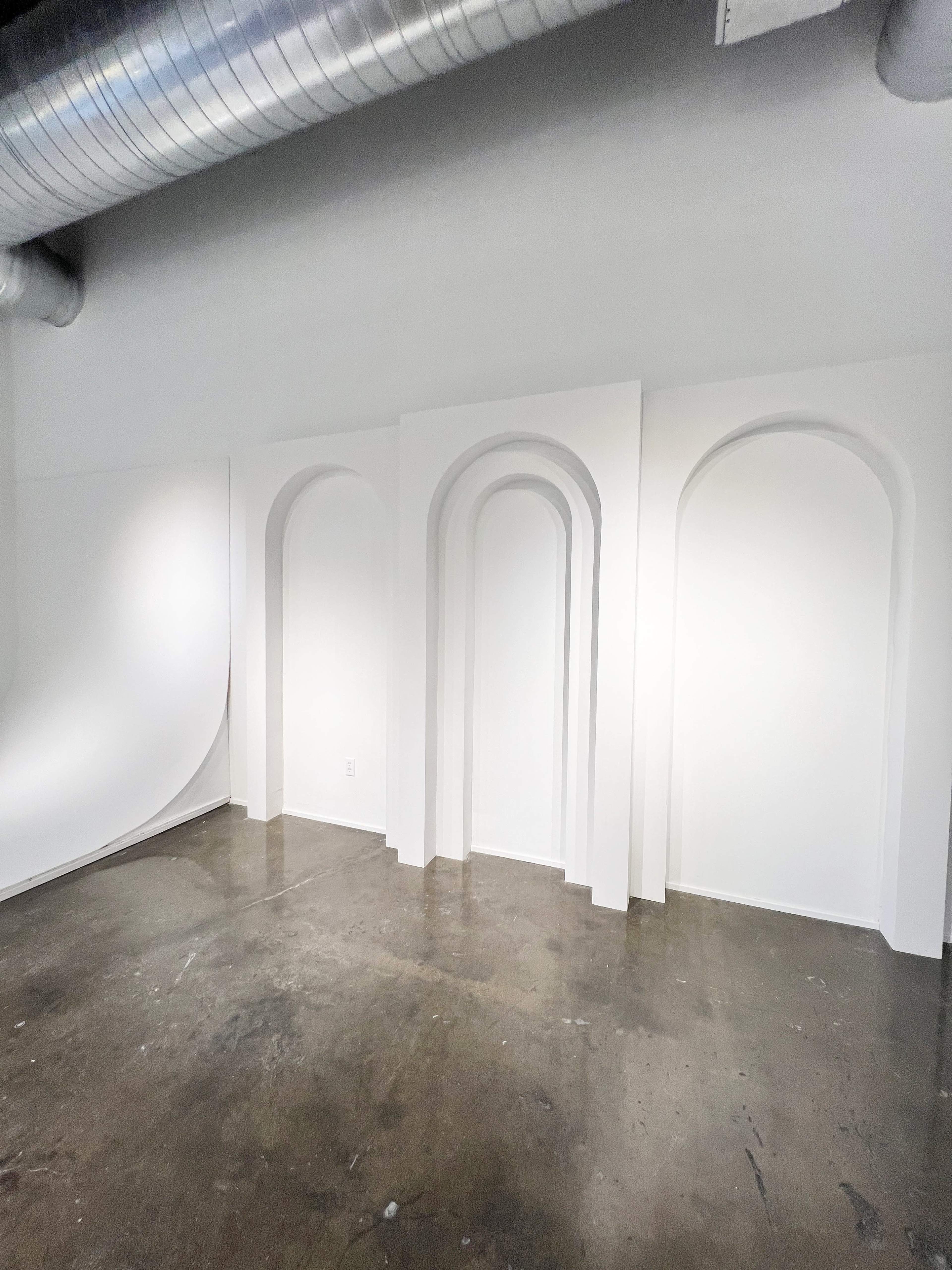 The image shows a minimalist interior space with three white arched recesses set against a light-colored wall.