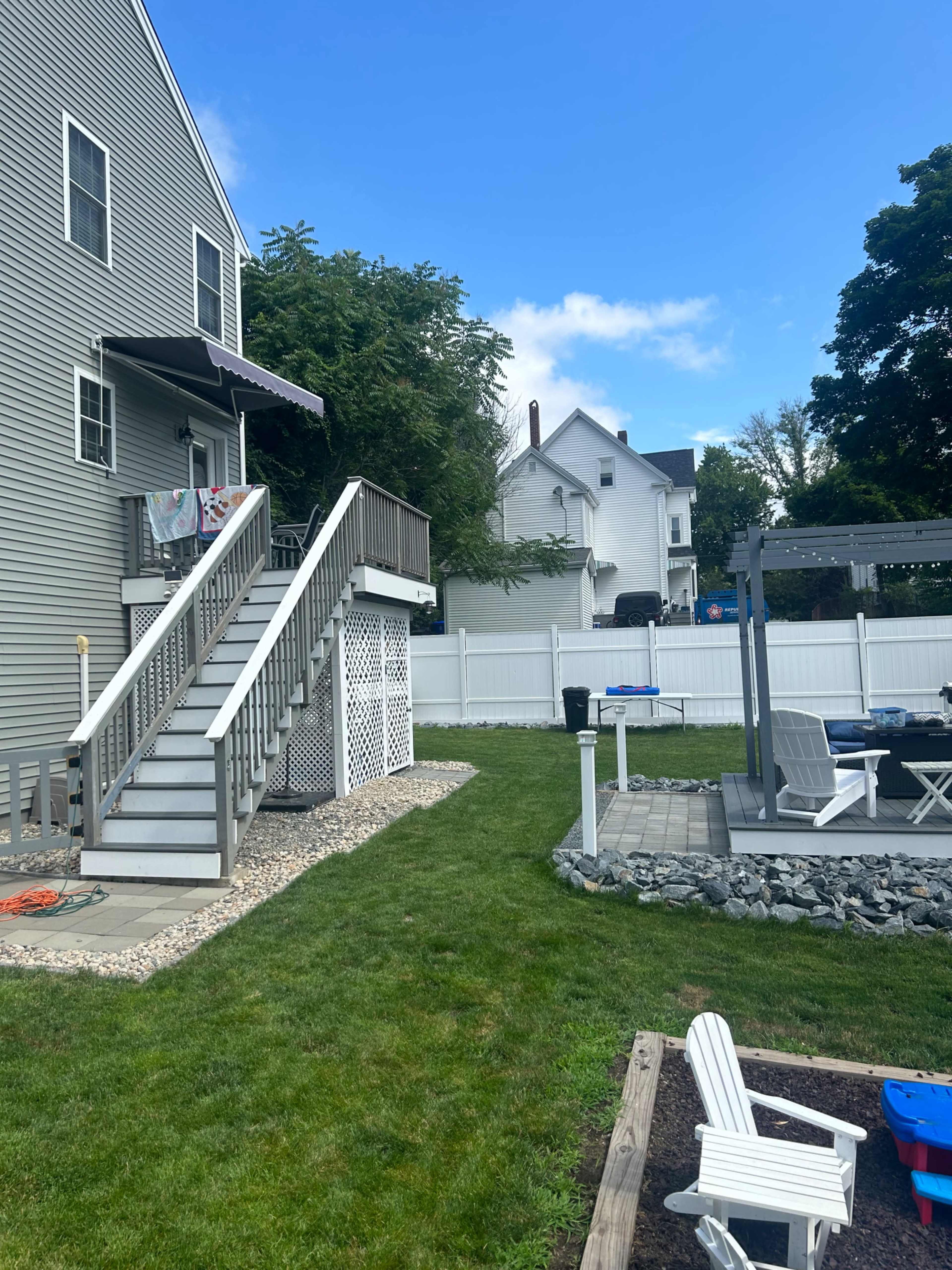 A two-story home with a wooden deck and staircase overlooks a grassy backyard, which features a stone patio and seating area.