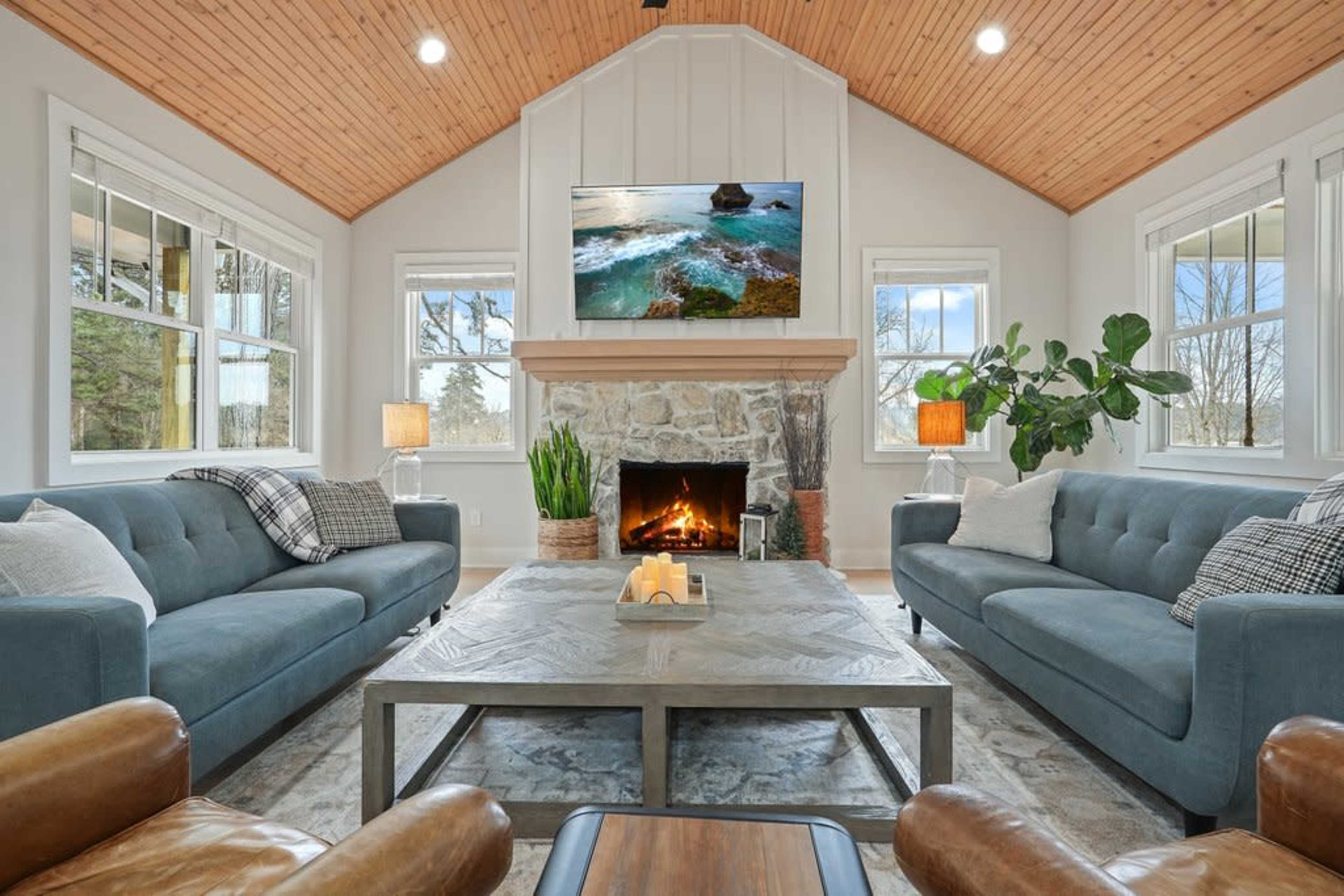 A cozy living room features two blue sofas facing a stone fireplace, with a wooden ceiling and a television mounted above the mantel.