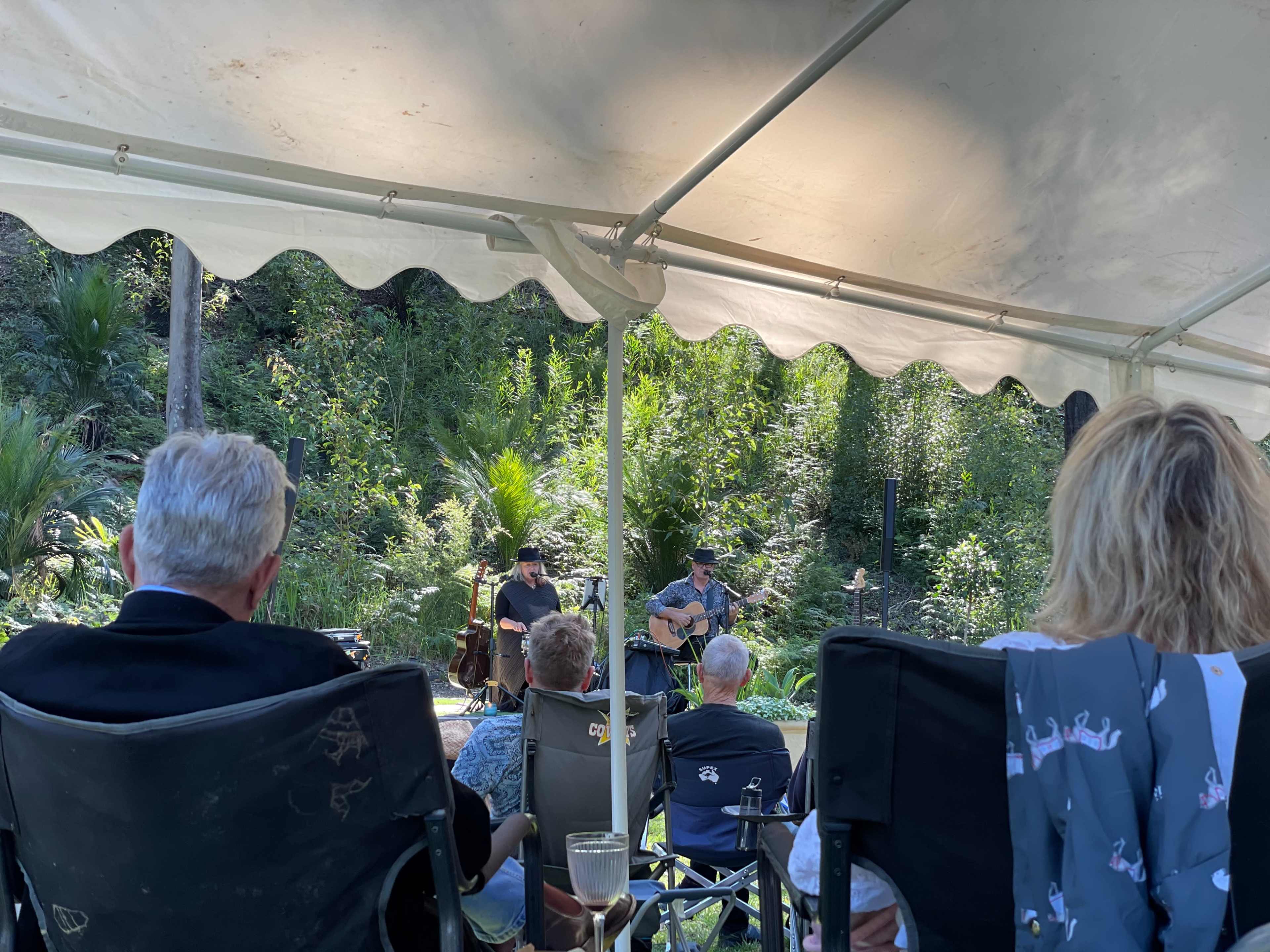 Two musicians perform on a small stage under a canopy, while an audience sits in folding chairs observing the performance amidst a wooded backdrop.