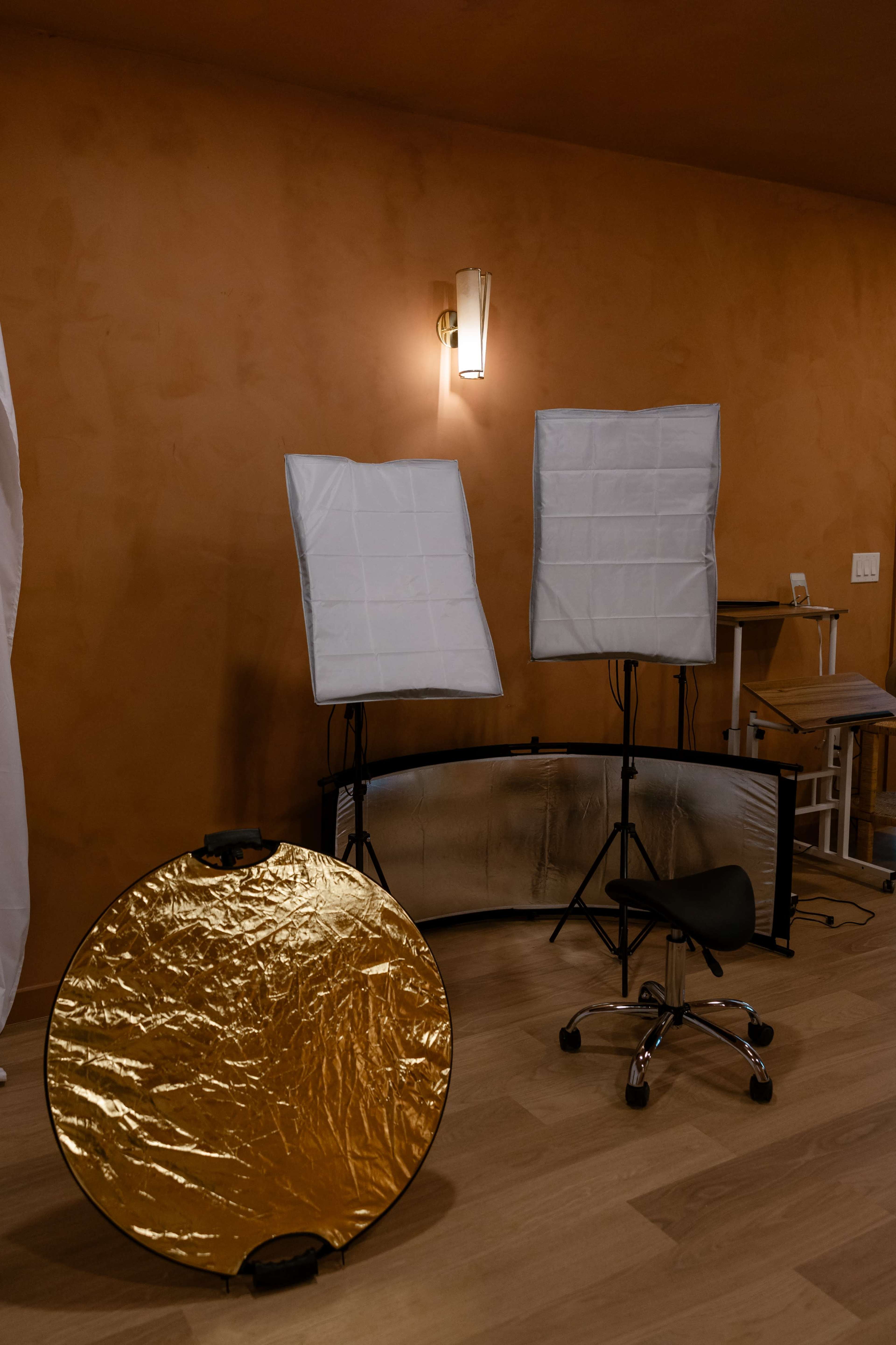 The image shows a photography studio setup with two softbox lights, a gold reflector disc on the floor, and a black swivel chair.