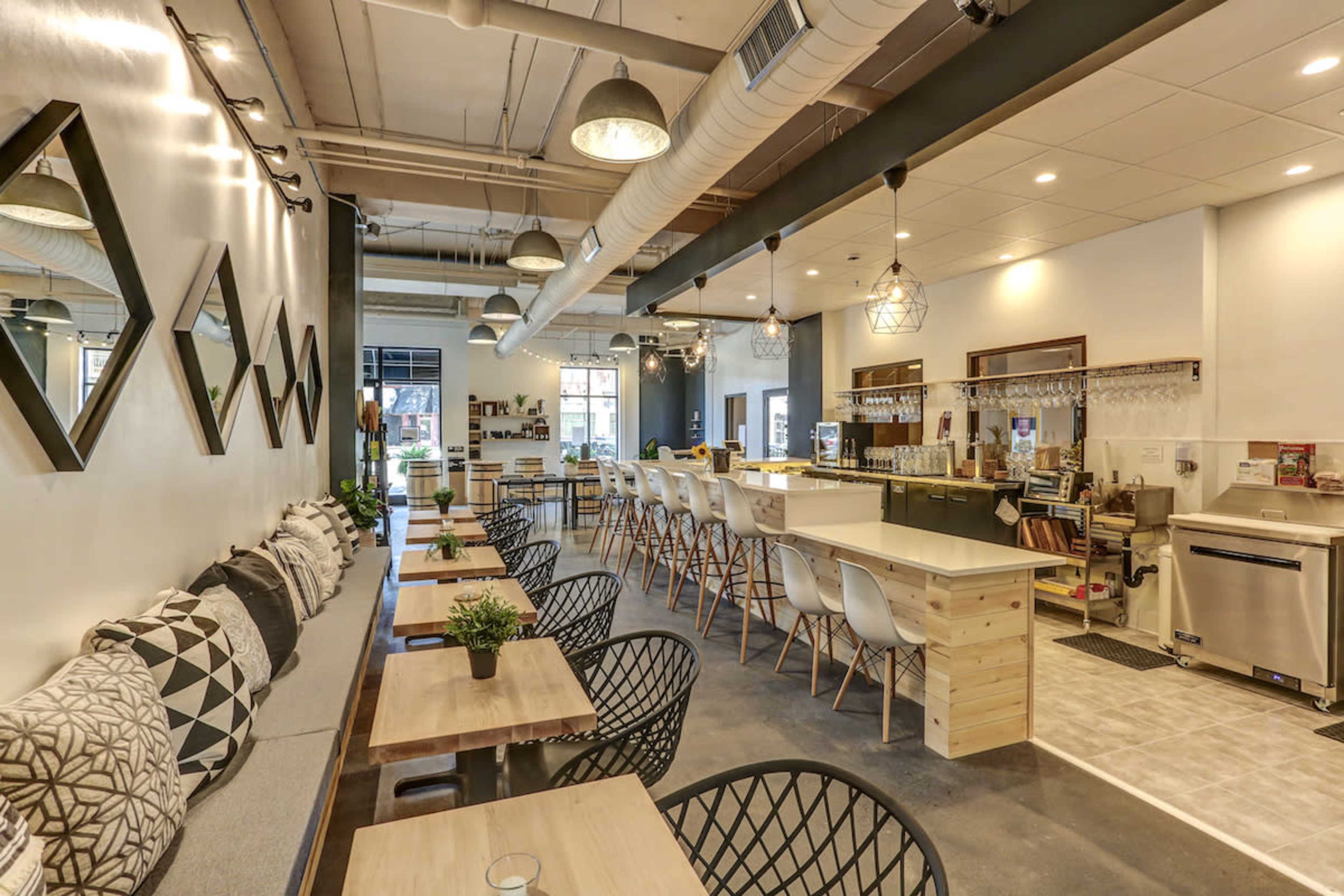 The image shows a modern café interior with a seating area featuring wooden tables, black chairs, and a long bar counter with stools.