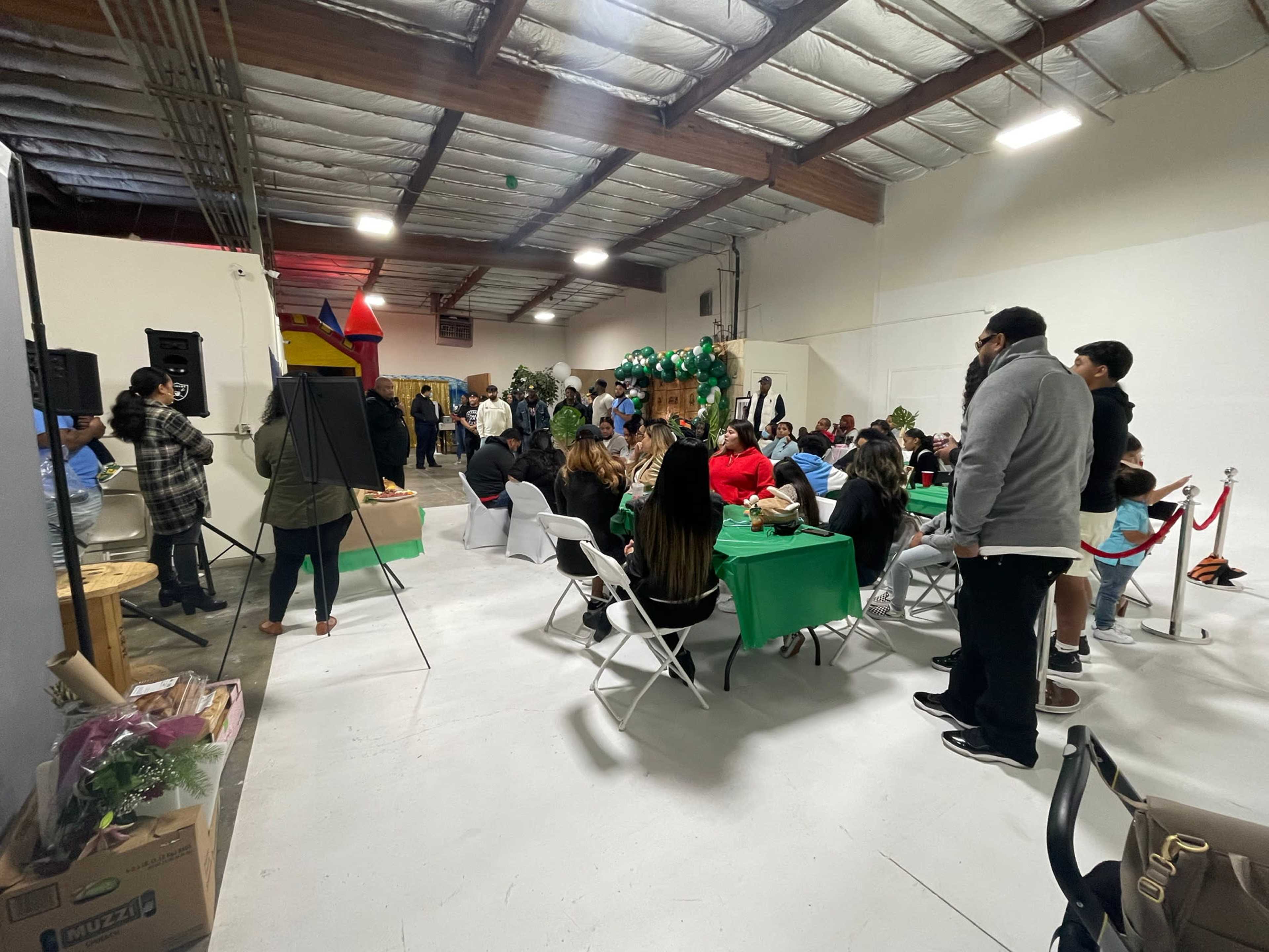 A crowd of people gathers around tables in a large indoor space decorated for an event, with some attendees interacting near a stage.