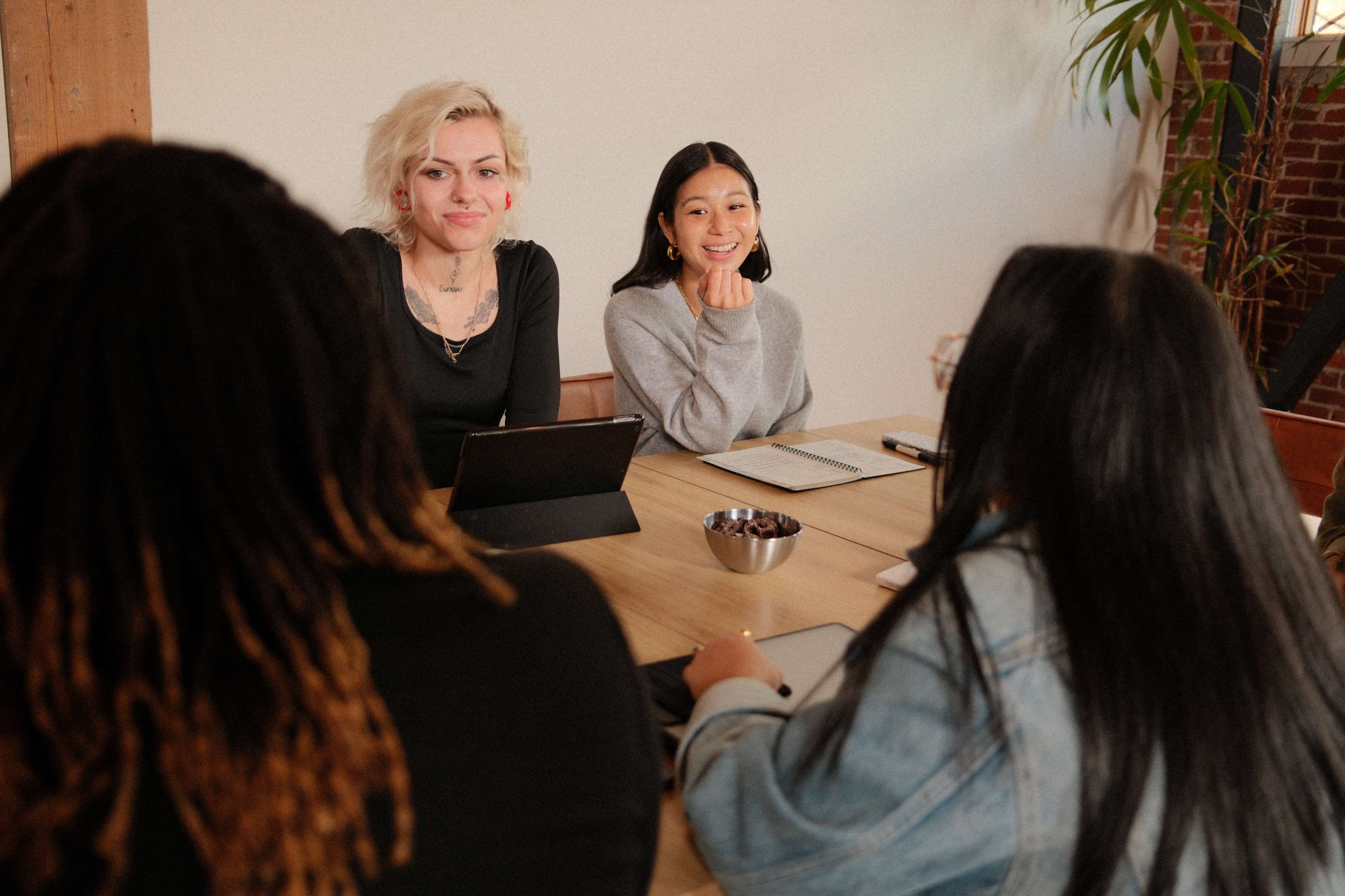 A group of five people sit around a wooden table in a meeting room, engaged in conversation, with some using tablets and notebooks.