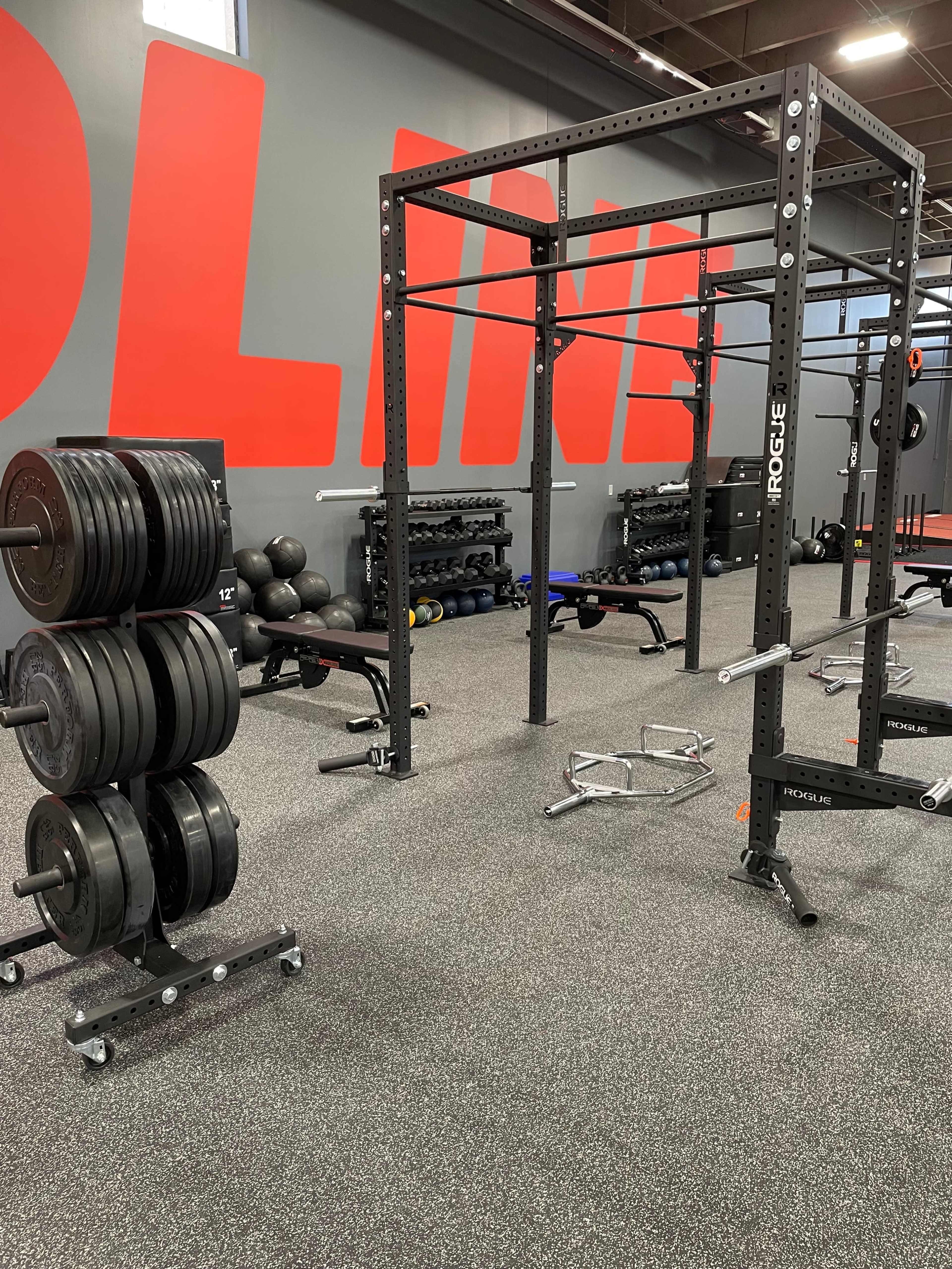 The image shows a gym interior featuring a weightlifting rack, a set of weight plates on a cart, and various exercise equipment against a wall with bold red letters.