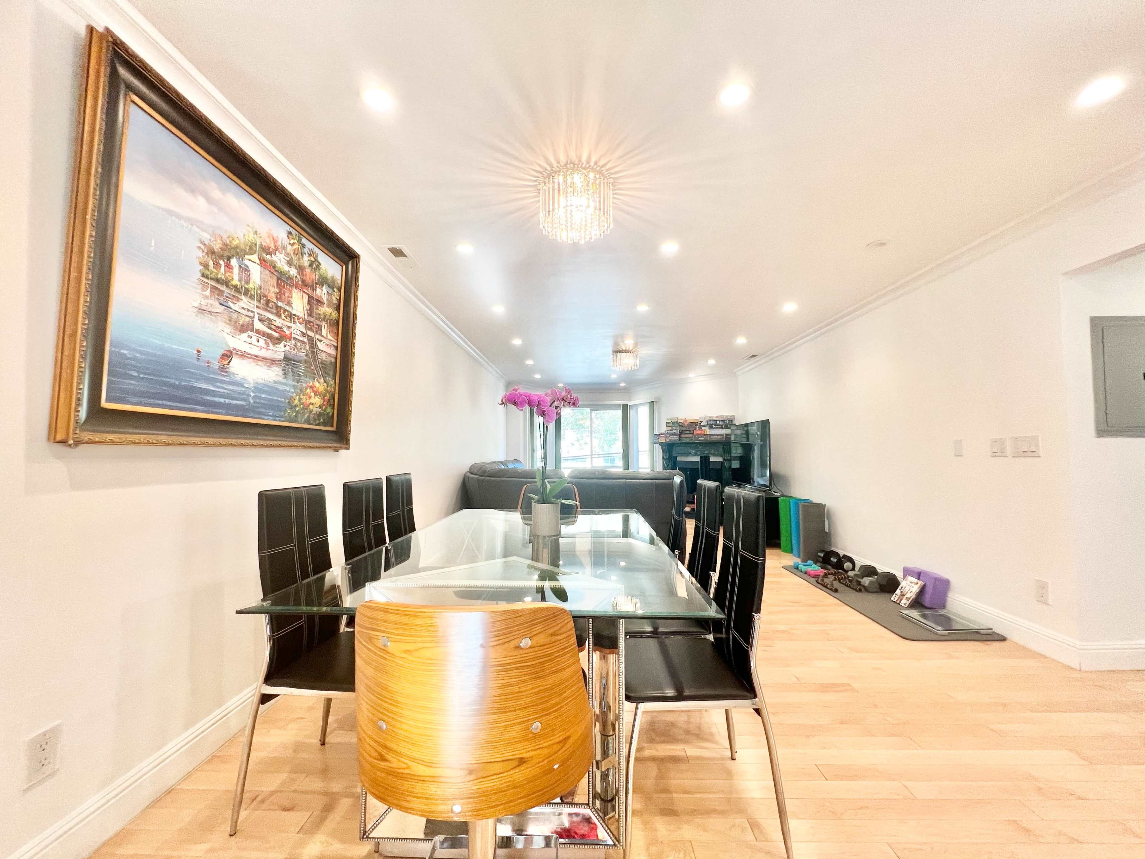 A modern dining area features a glass table surrounded by black chairs, with a painting on the wall and a view towards a living space equipped with exercise equipment.