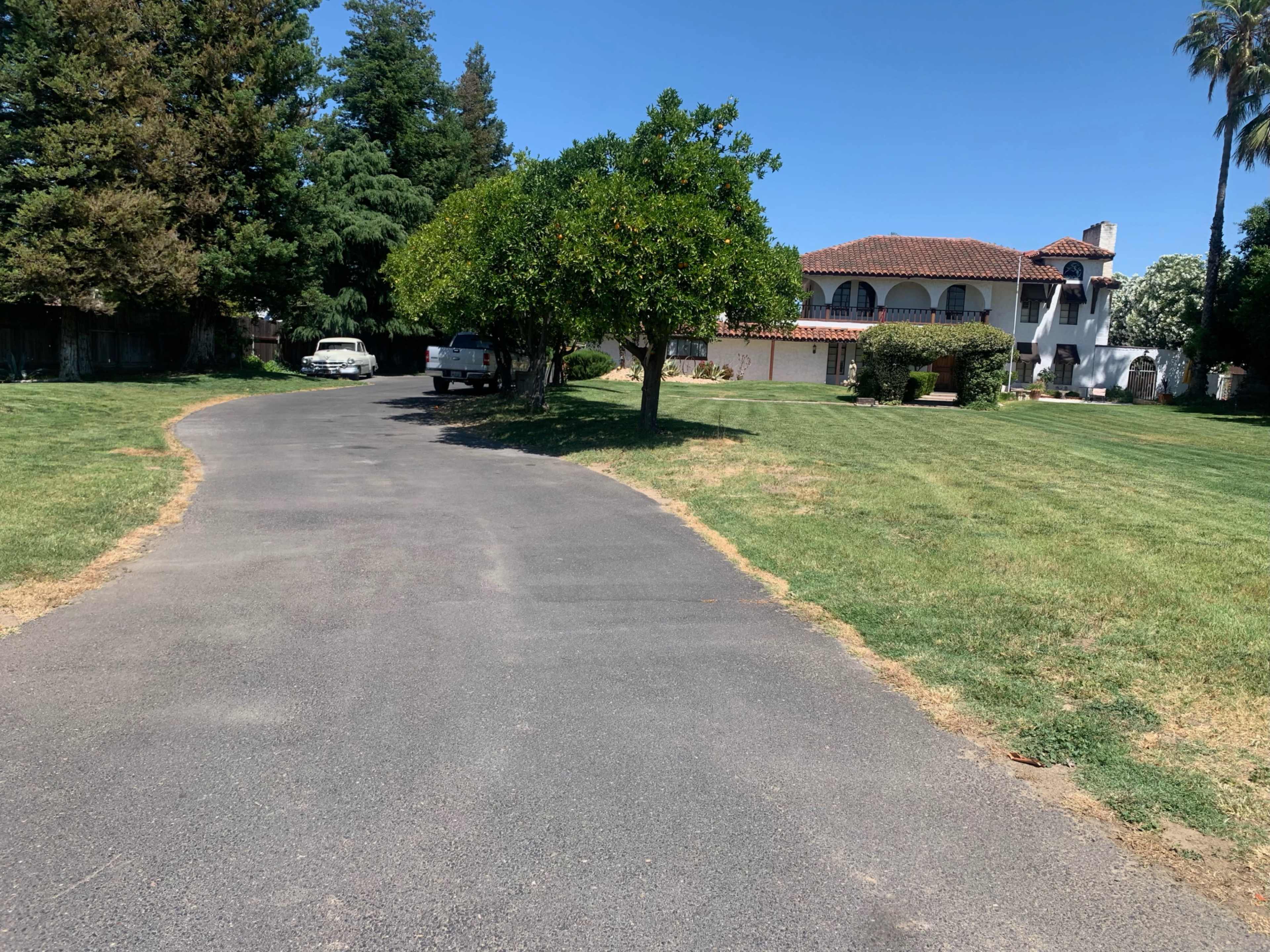 A paved road leads to a two-story house with a red tile roof surrounded by trees and a grassy lawn.