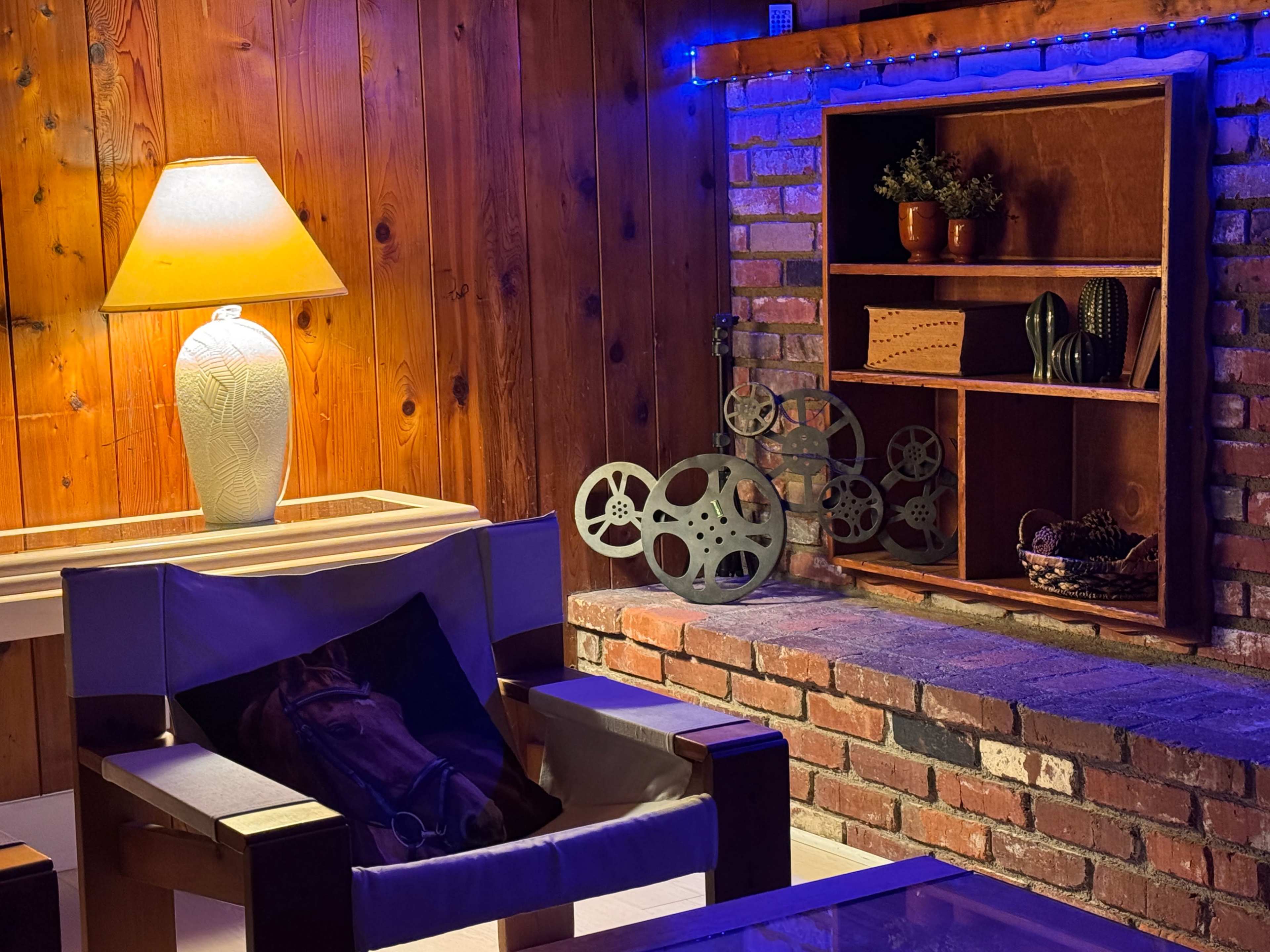 The image shows a cozy interior with wooden walls, a lamp, a comfortable chair, a brick fireplace, and a display shelf containing decorative items.