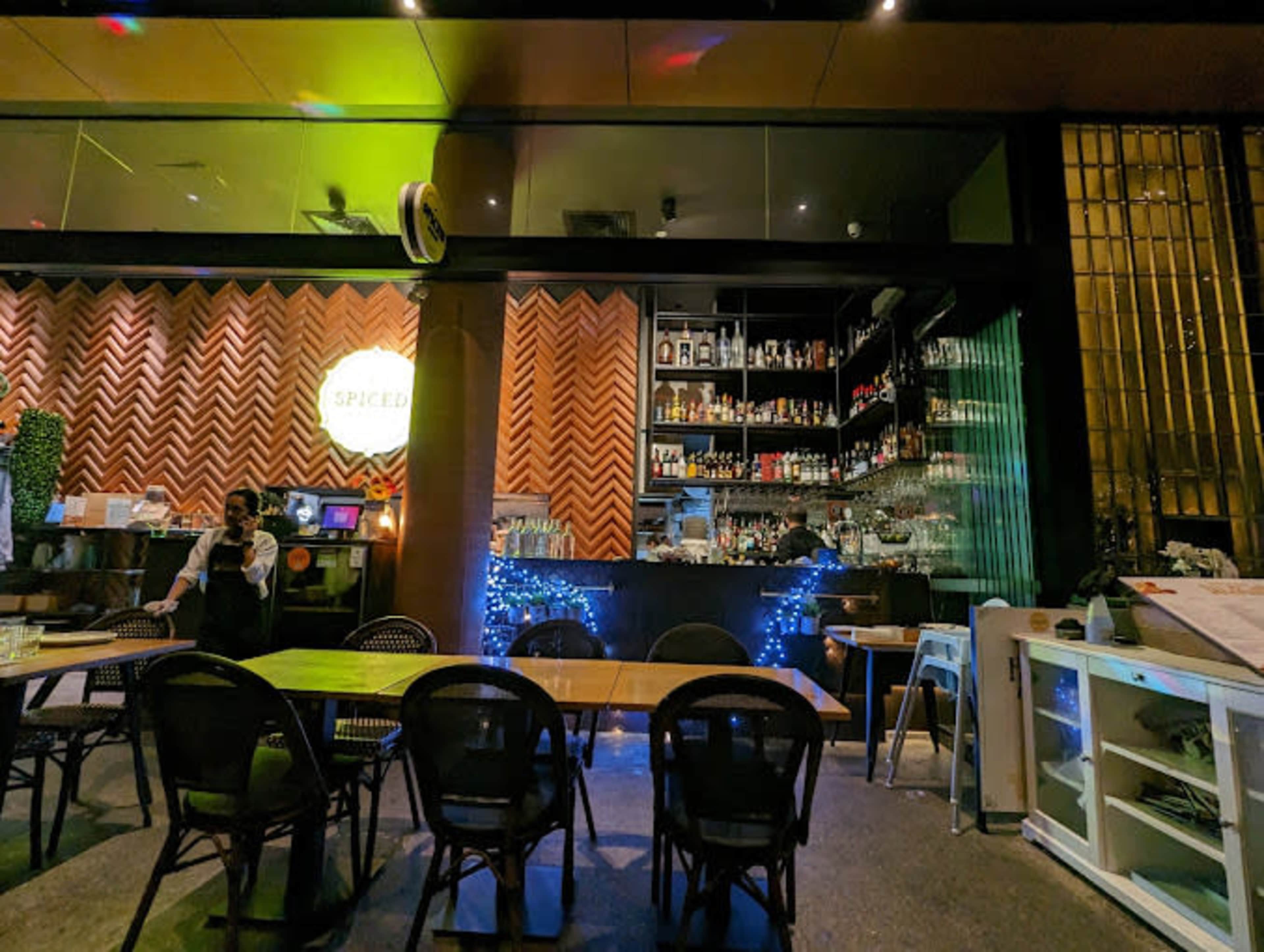 The image shows the interior of a bar or restaurant with a wooden wall, a well-stocked shelf of bottles, and tables arranged for dining.