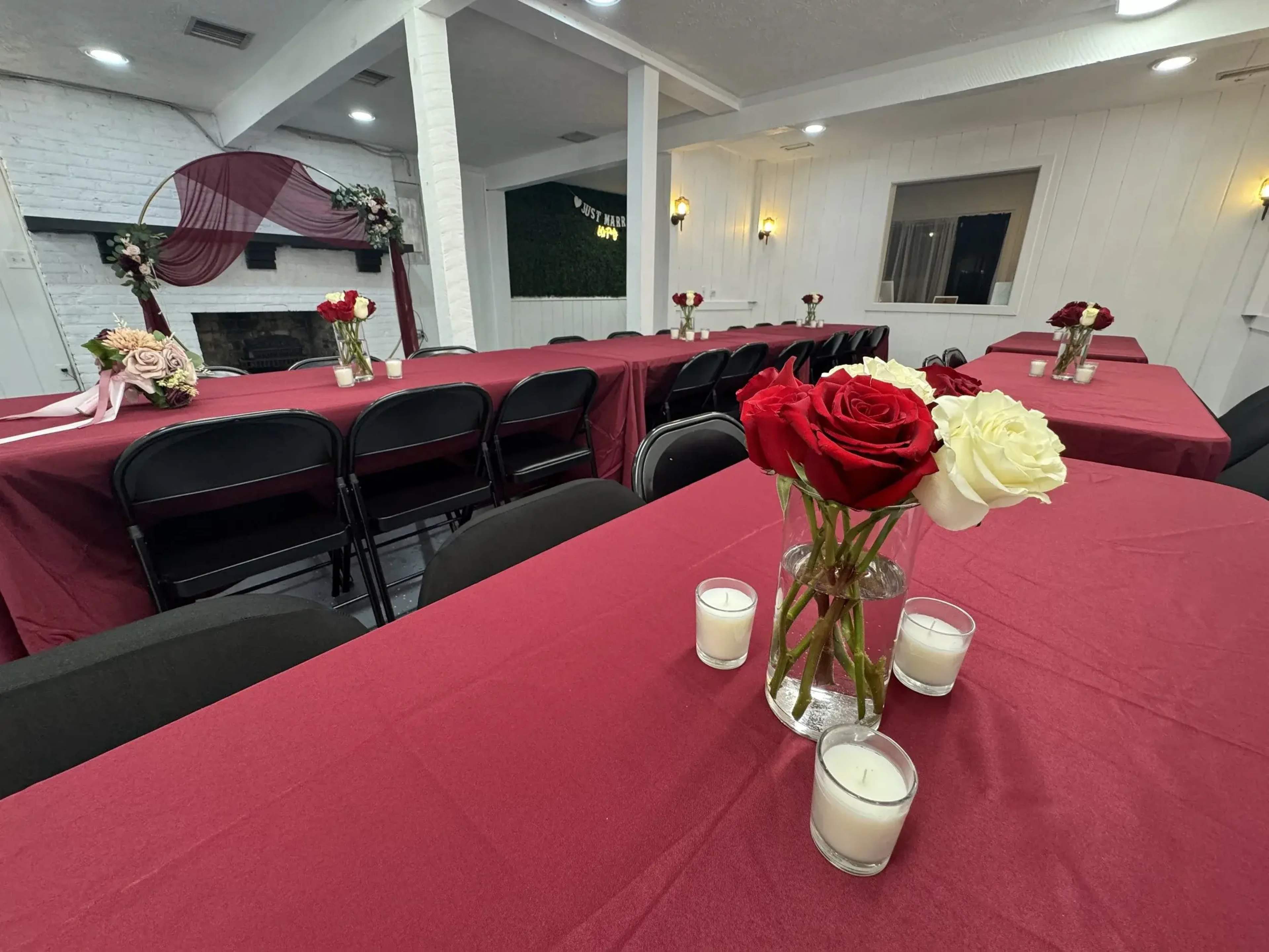 The image shows a decorative space set up for an event, featuring tables covered in burgundy tablecloths, black chairs, and centerpieces of red and white roses in glass vases alongside candles.