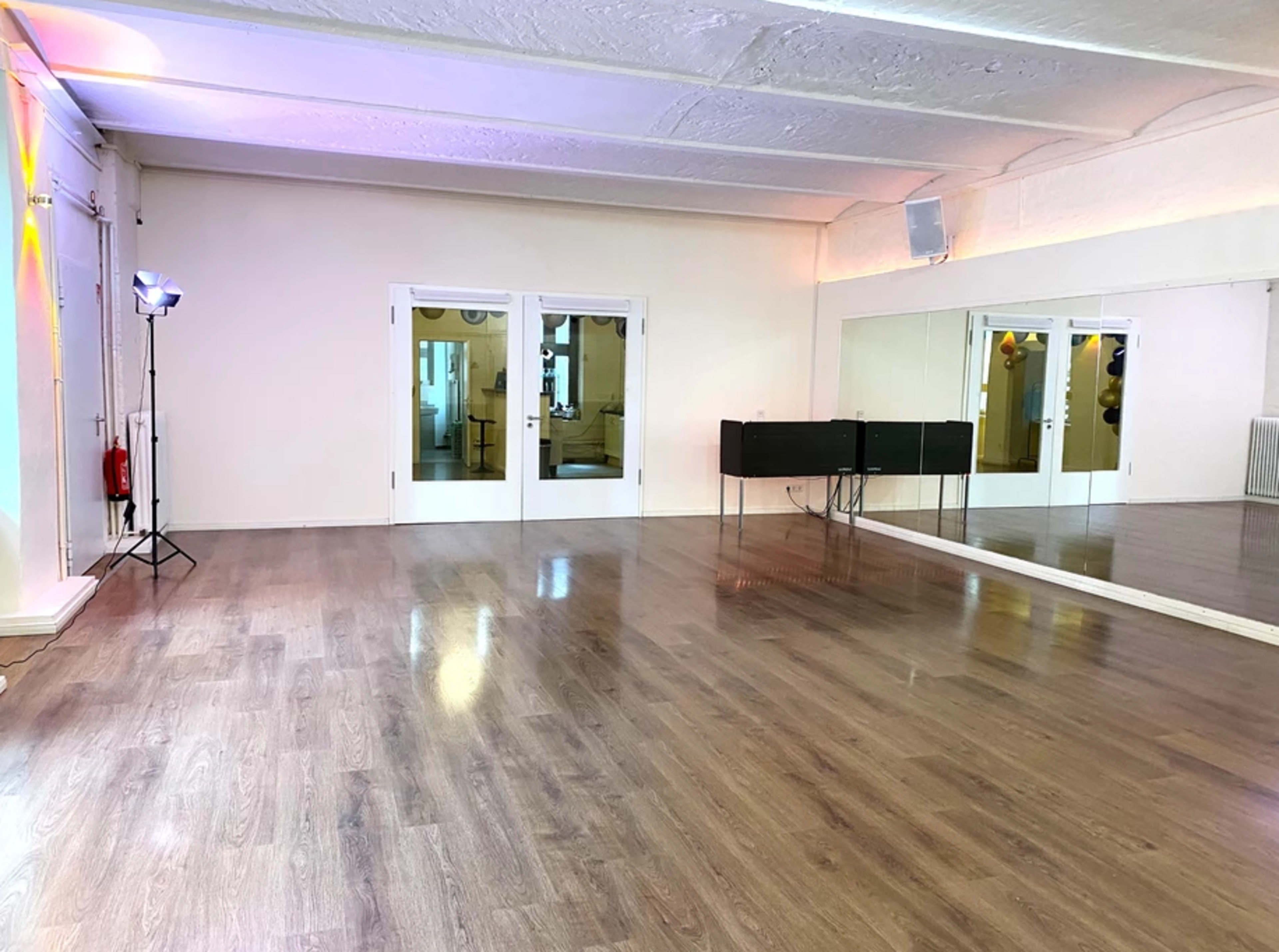 An empty dance studio with a wooden floor, large mirrors on one wall, and light fixtures in the ceiling.