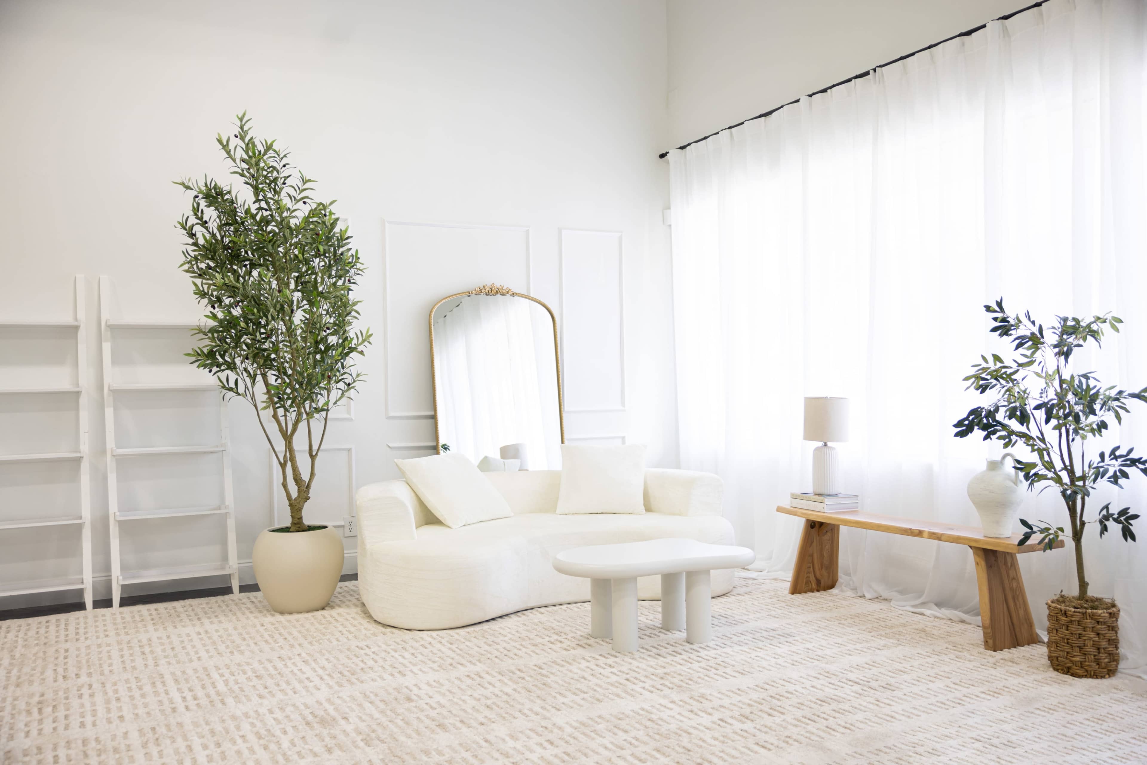The image shows a modern, minimalist living room with a white couch, a round coffee table, a wooden bench, and a tall plant next to a mirror and shelves.