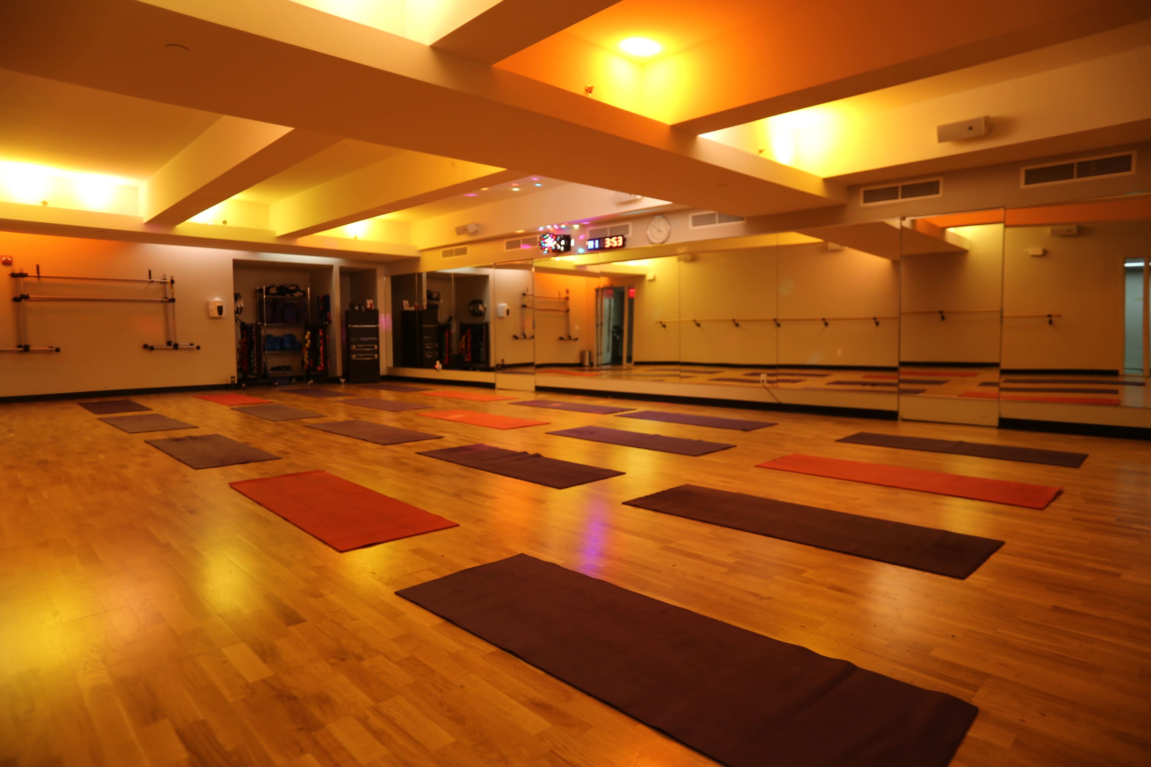The image shows a spacious yoga studio with wooden flooring, multiple exercise mats arranged on the ground, and mirrors lining one wall.
