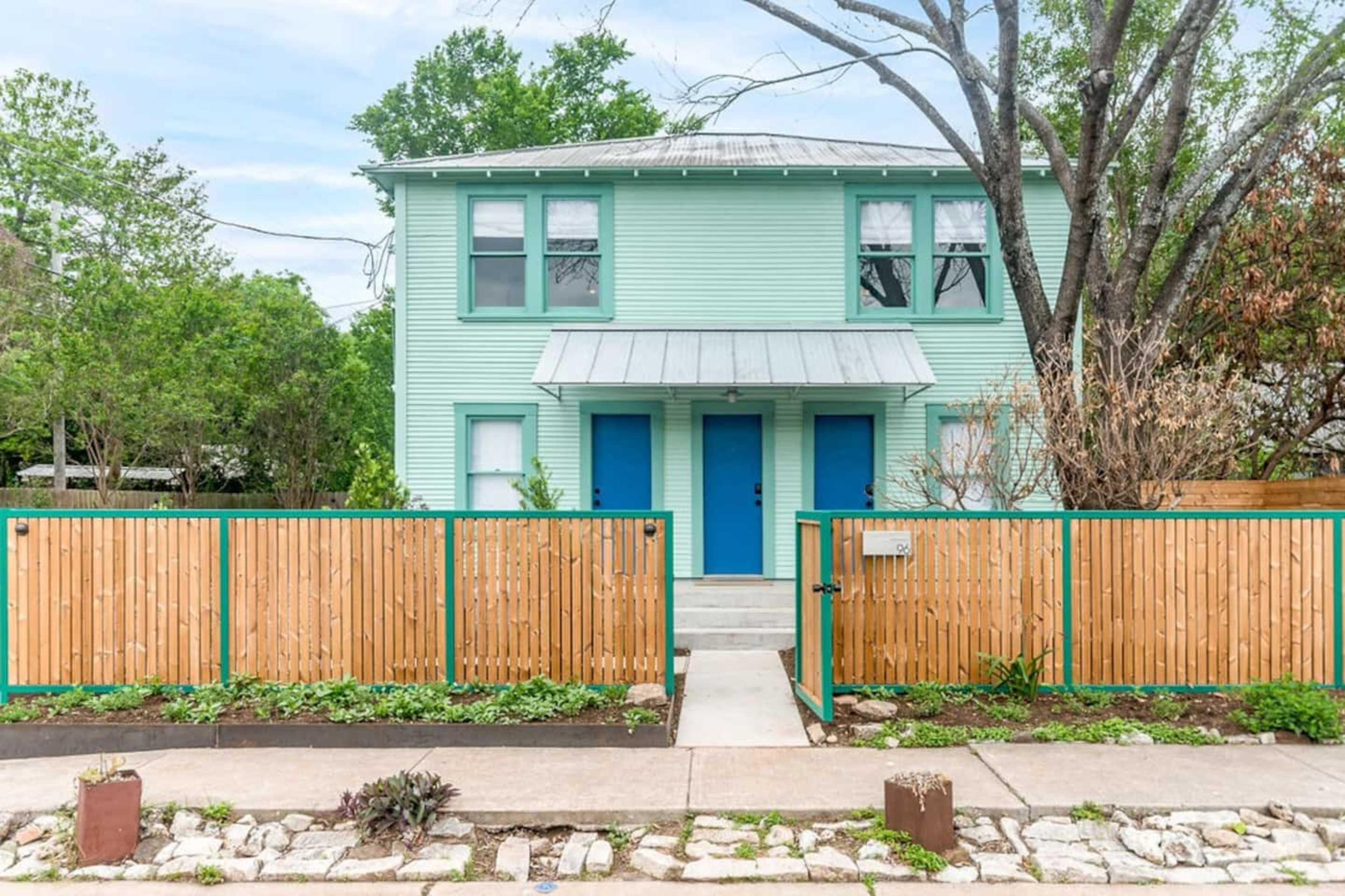 A two-story, mint green duplex with blue doors and a fenced front yard is shown.