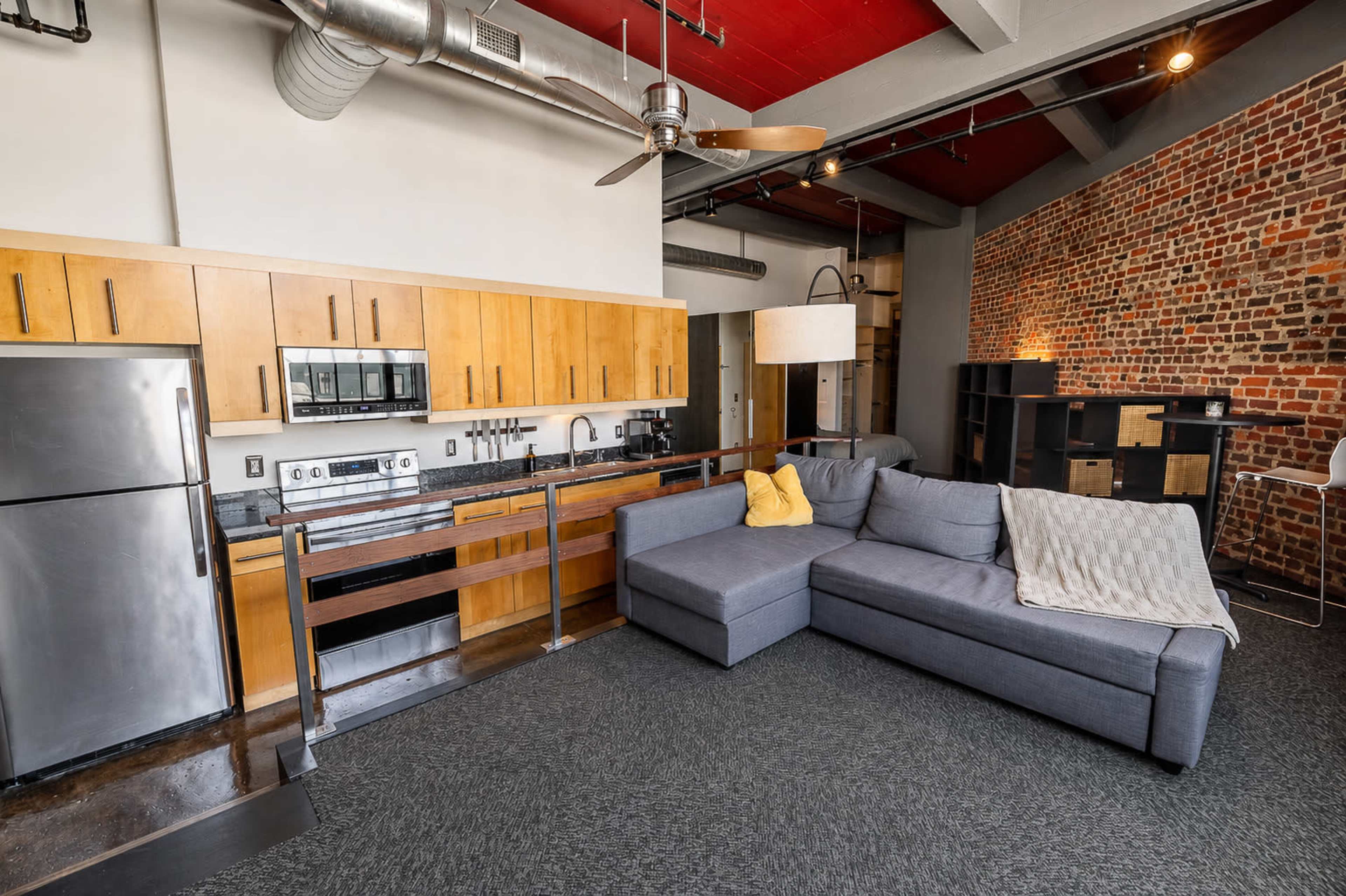 The image shows a modern living space with a sectional sofa, open kitchen area featuring wooden cabinets, stainless steel appliances, and a brick accent wall.