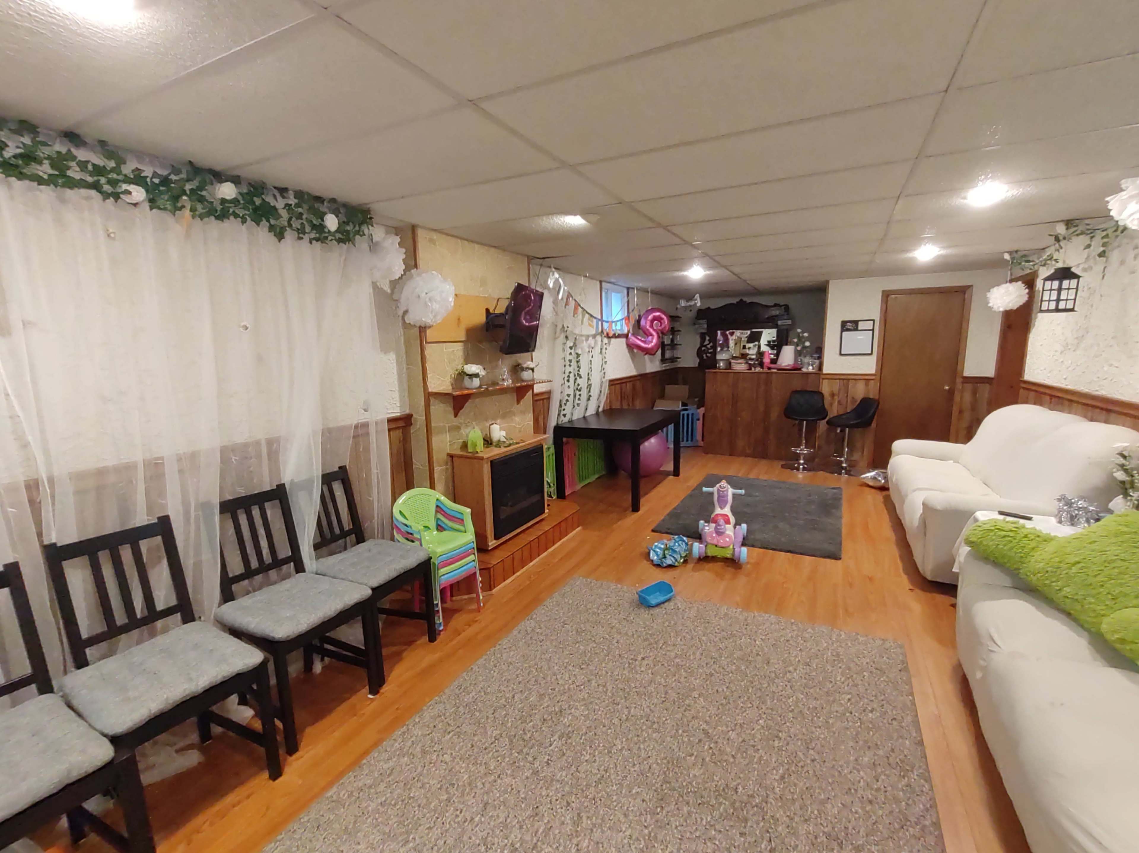 A basement living area features a set of chairs along one wall, a couch, a small table, a bar area, and a play space with scattered toys.