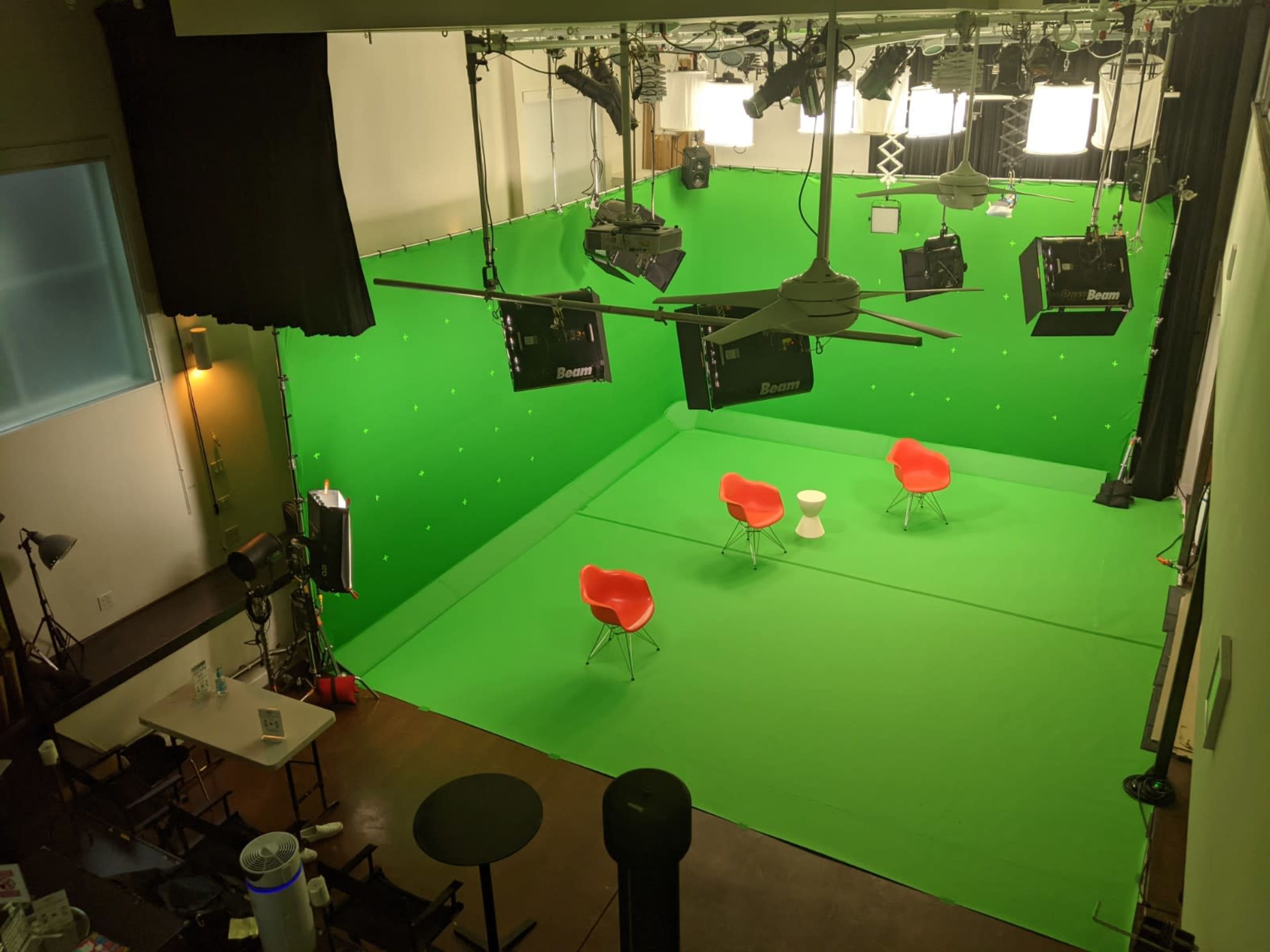 The image shows a studio with a green screen backdrop, featuring three red chairs arranged around a small white table, along with studio lighting equipment overhead.