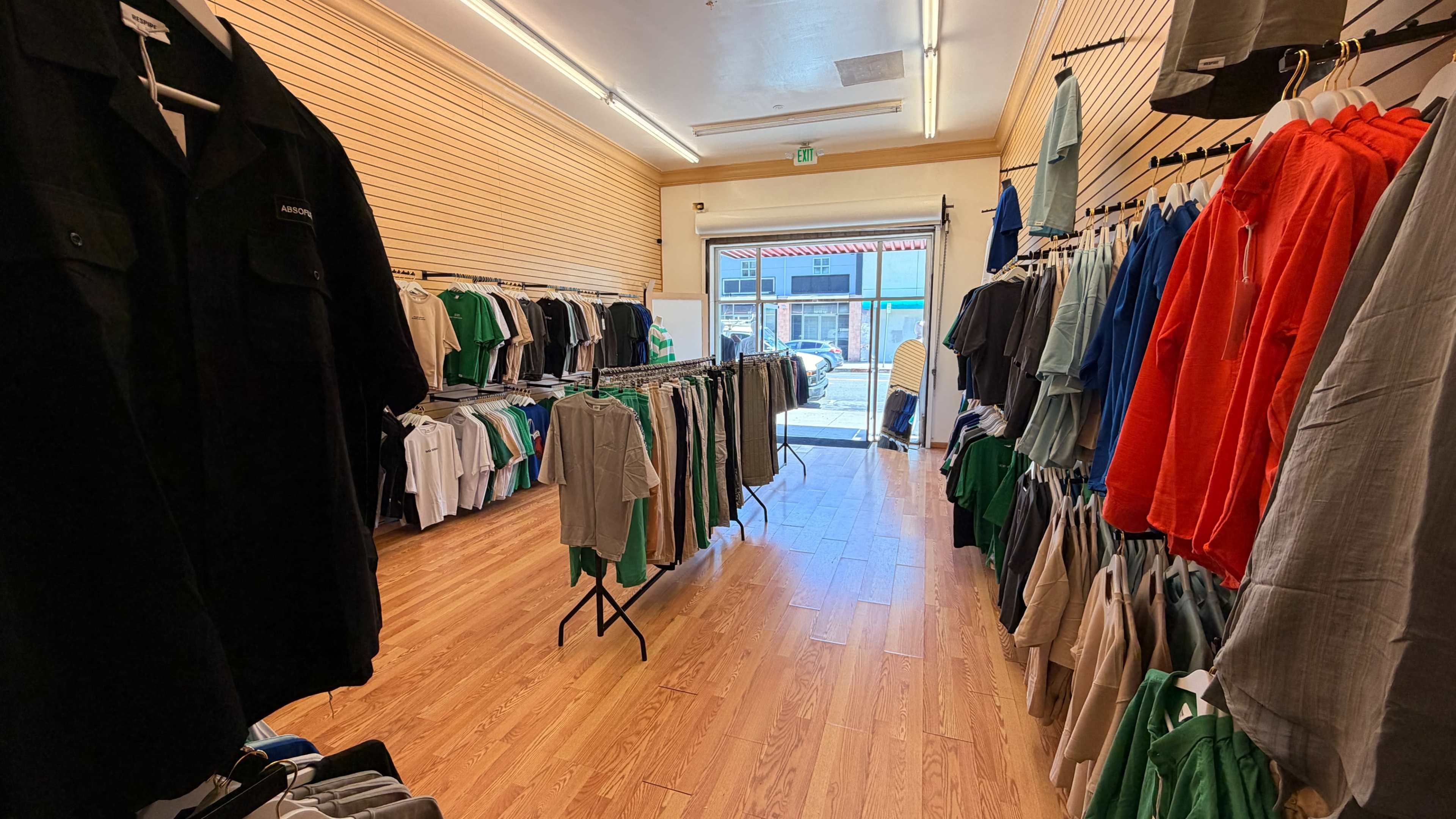 The image shows the interior of a clothing store with racks displaying various shirts and pants, and a door leading outside.