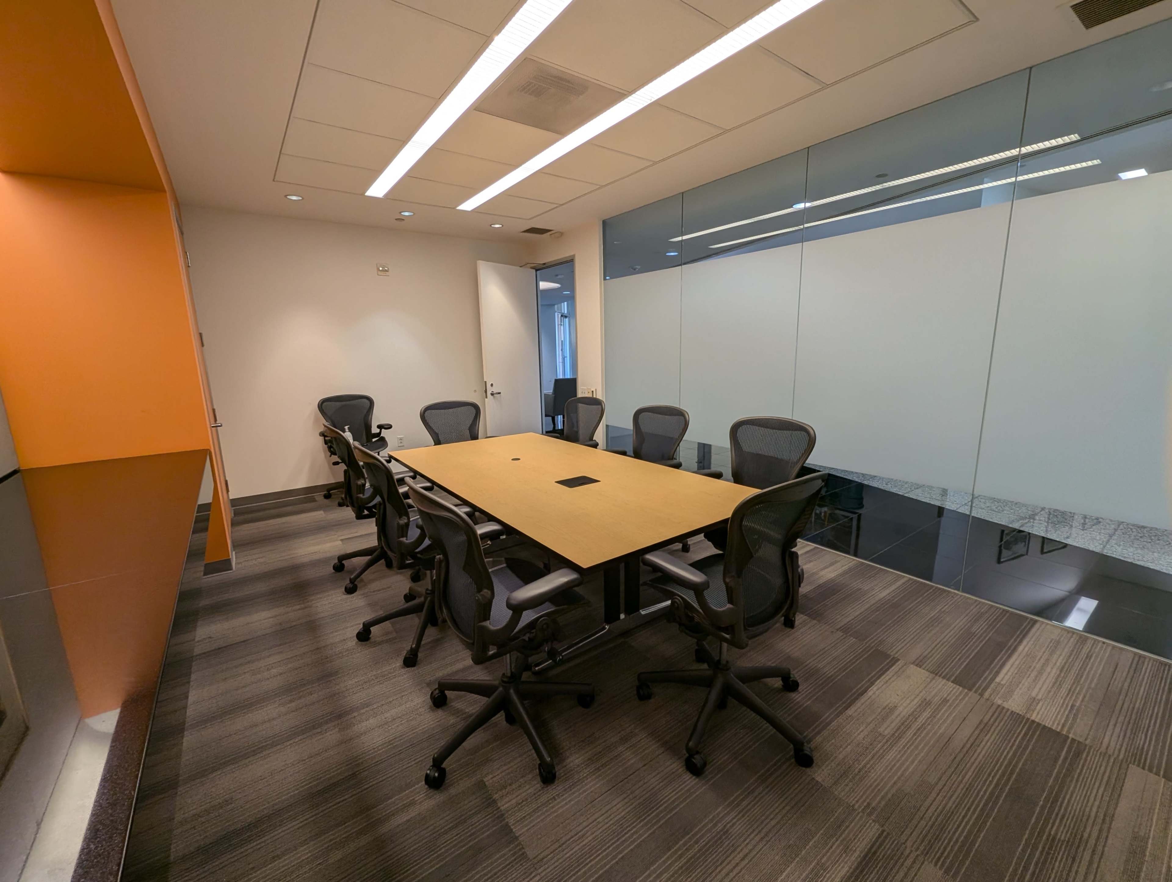 A conference room features a central table surrounded by six ergonomic chairs, with glass walls and modern lighting.