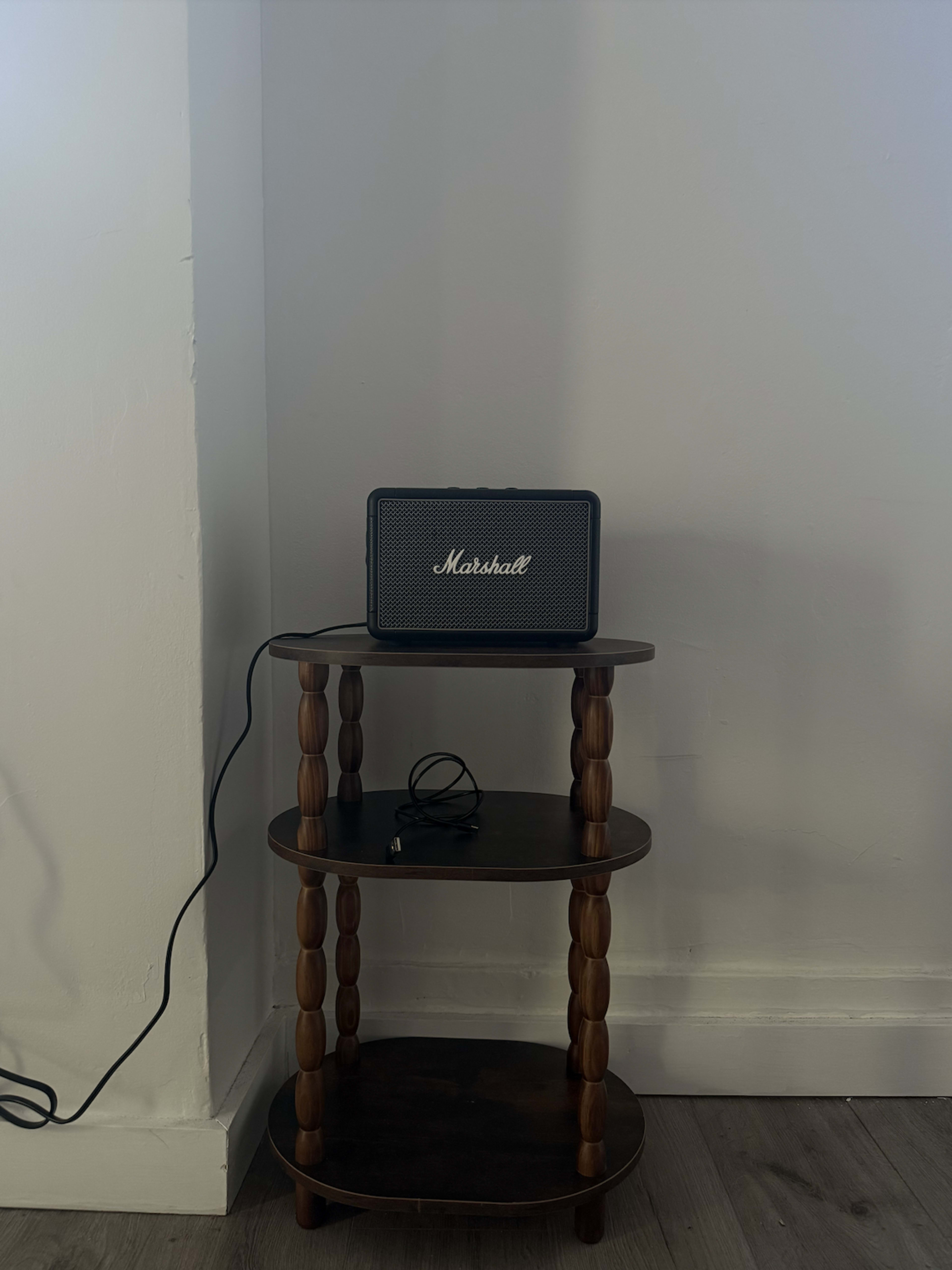 A Marshall speaker is placed on a two-tiered wooden table against a plain white wall.