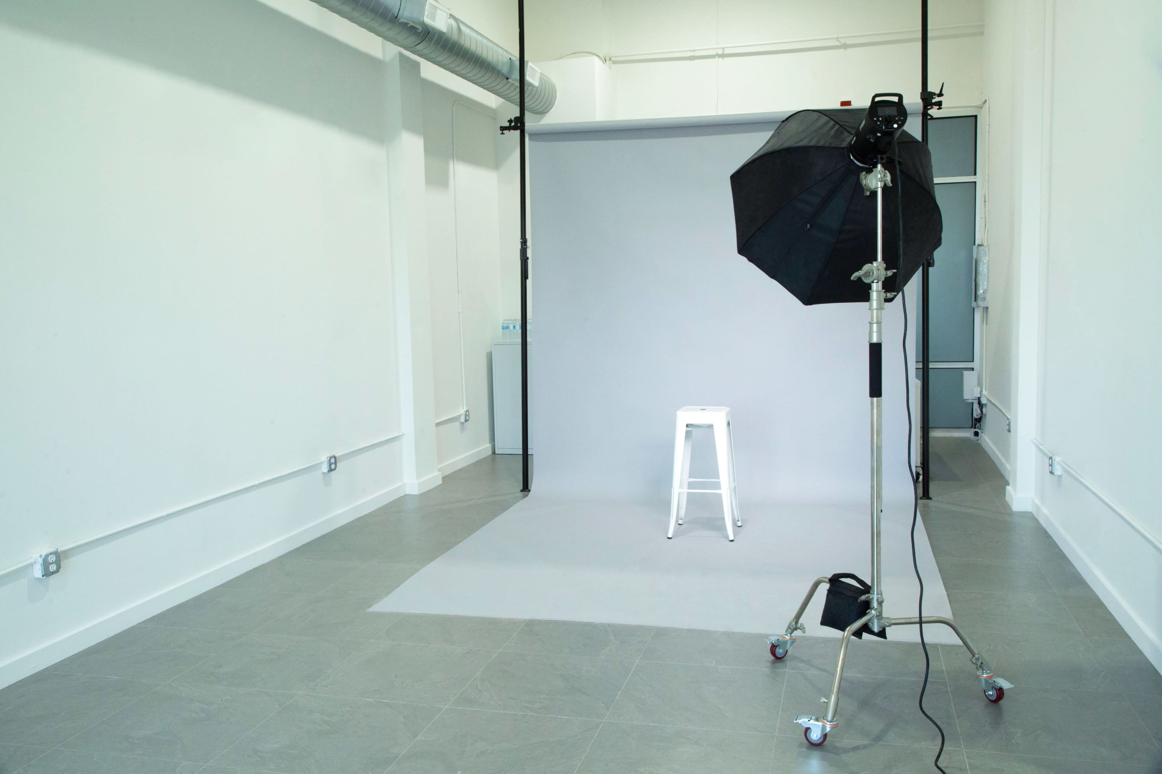 A photography studio with a gray backdrop, a white stool, and a softbox light setup on a stand.