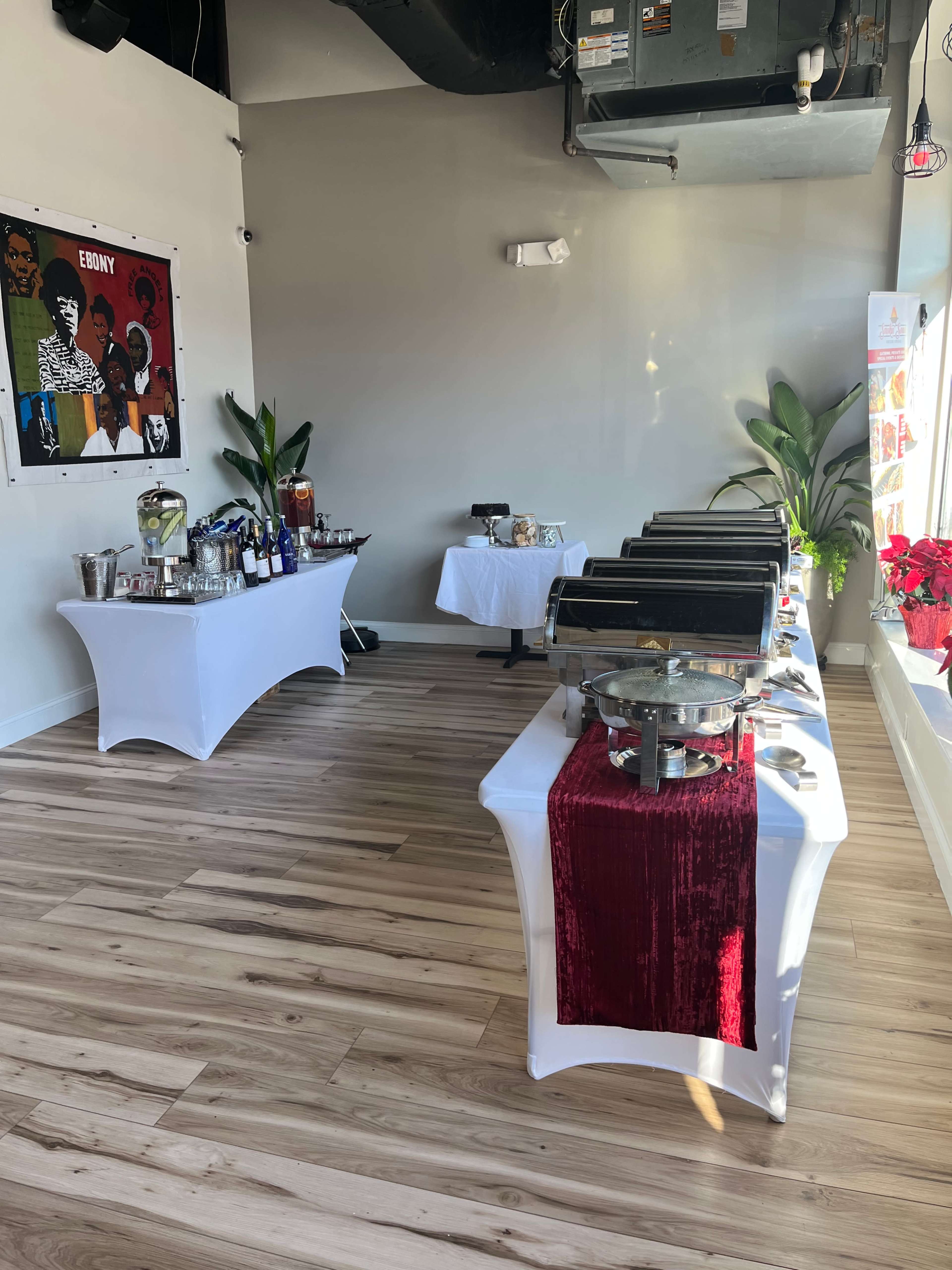 The image shows a banquet setup with several food trays on a long table, a smaller table with beverages, and decorations on the walls in a well-lit room.