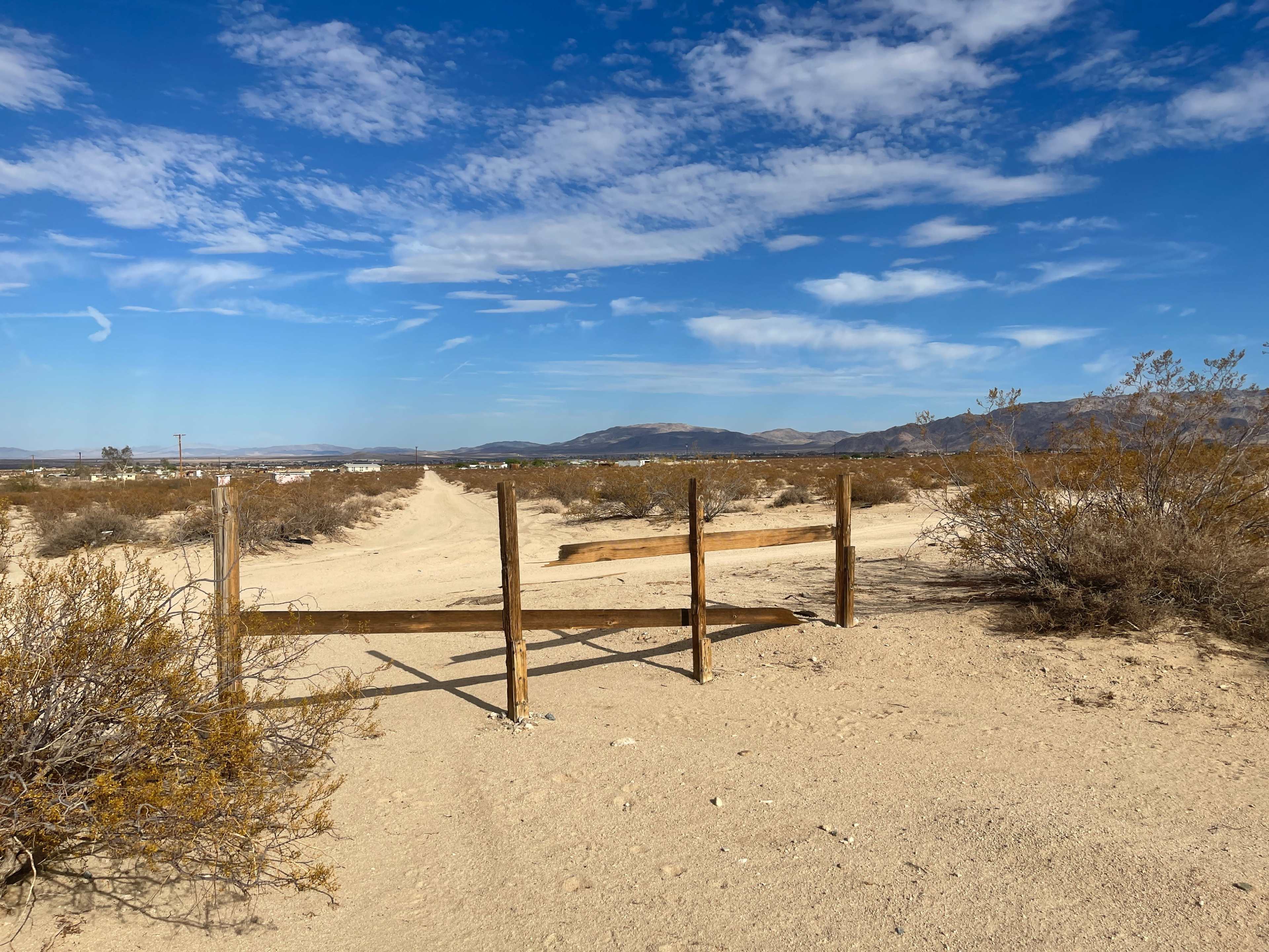 A dirt road leads through a dry landscape, marked by a wooden gate and surrounded by sparse vegetation under a blue sky with scattered clouds.