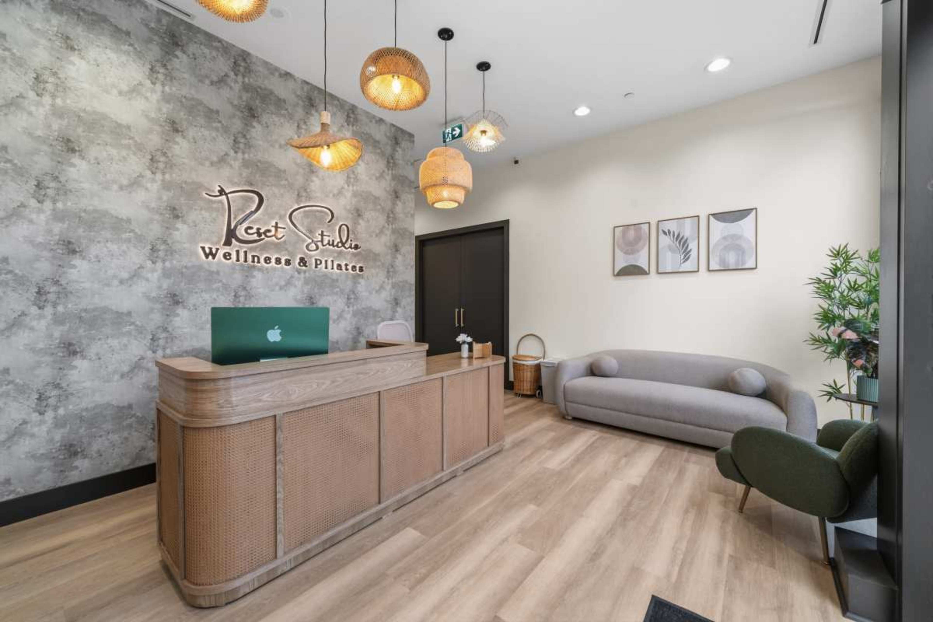 The image shows a wellness and Pilates studio reception area with a wooden front desk, decorative pendant lights, and modern furniture along with wall art.