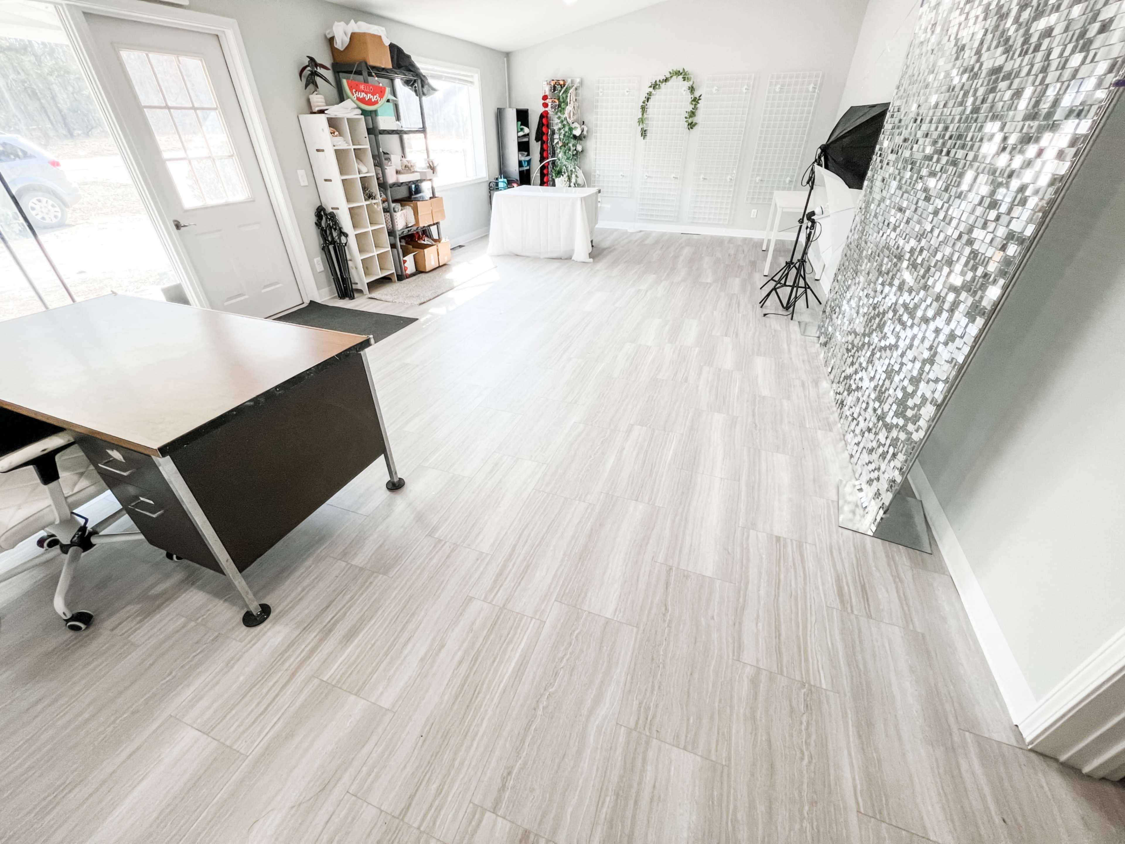 A spacious room with light-colored tiled flooring, a desk on wheels, and a mirrored wall, along with a white table and various props in the background.