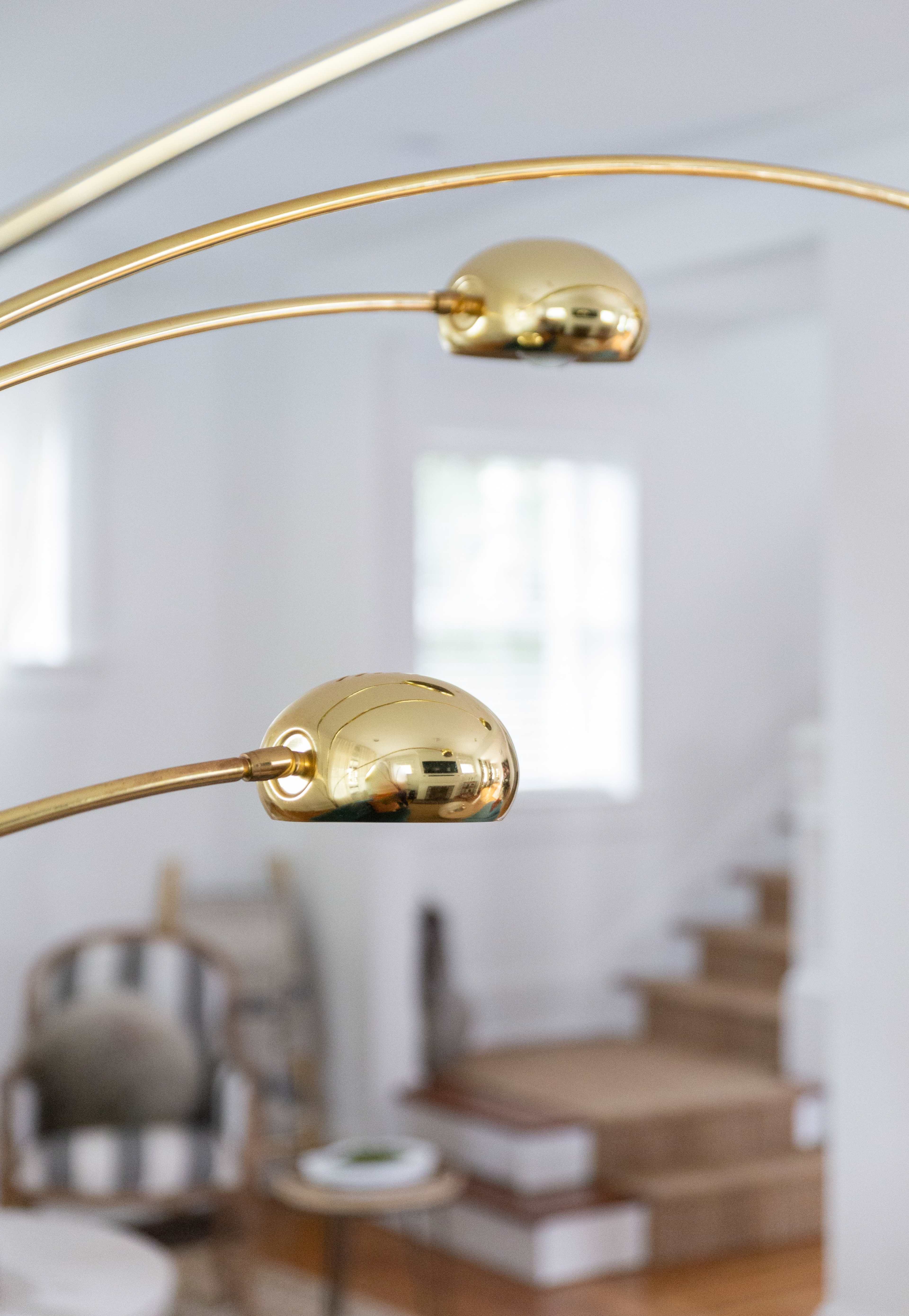 A close-up of a modern floor lamp with two curved gold lights, positioned in a brightly lit room with stairs in the background.