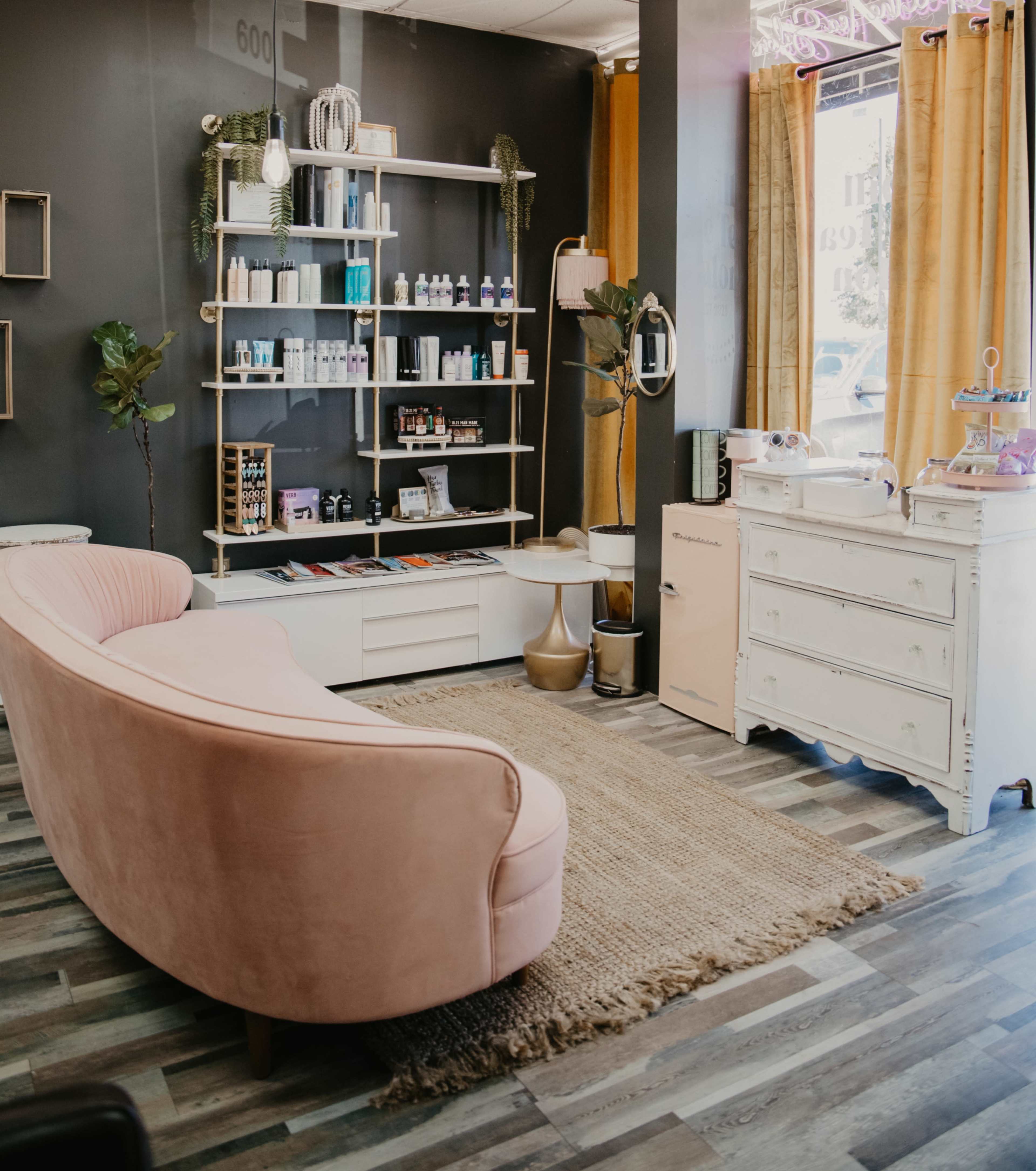 A modern beauty salon interior featuring a pink curved sofa, shelves with various beauty products, and a vintage-style dresser against a dark wall.