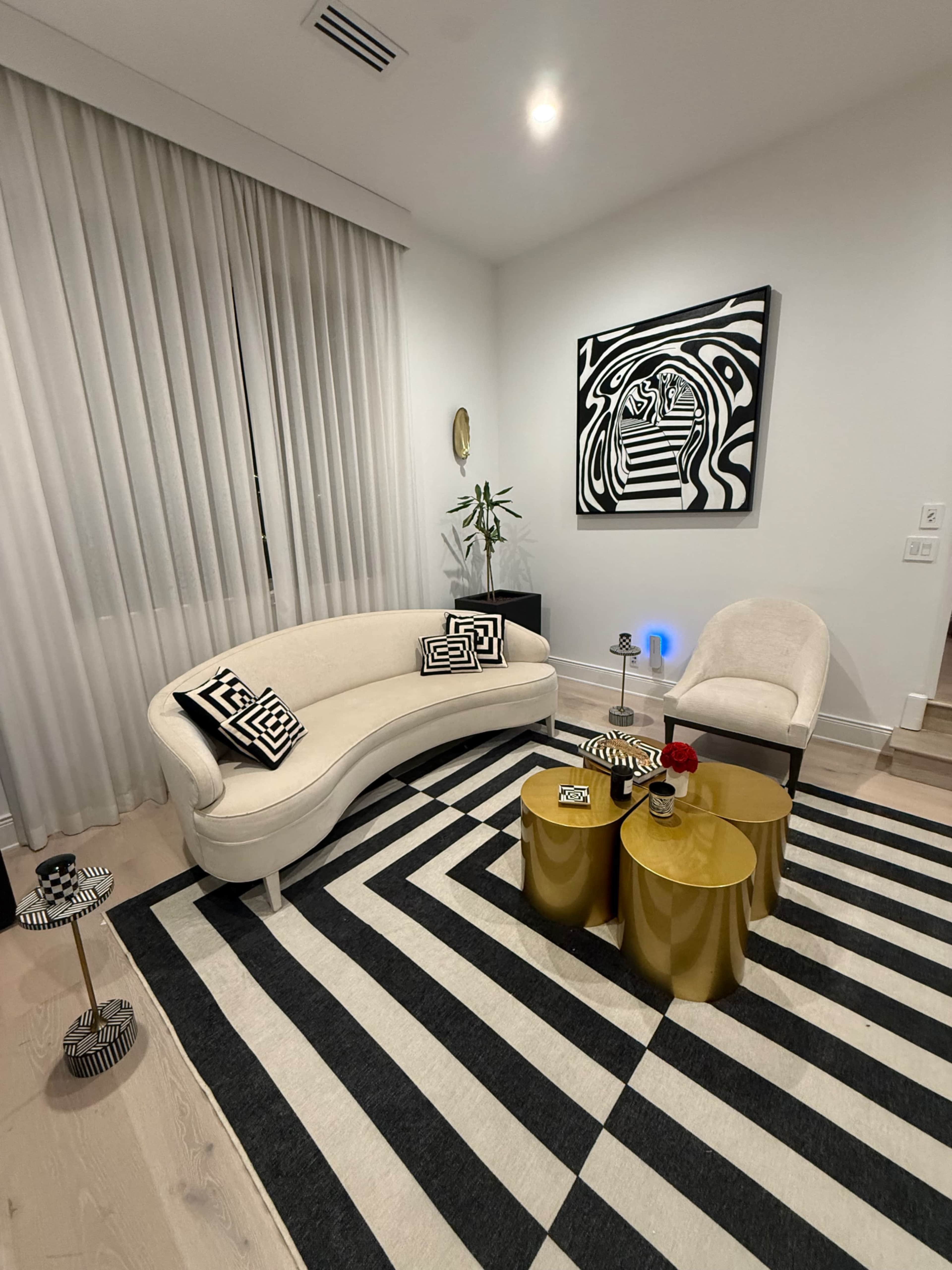 The image depicts a modern living room featuring a curved white sofa, a light-colored armchair, and two gold side tables on a black and white striped rug, with large windows covered by sheer curtains and a piece of abstract wall art.